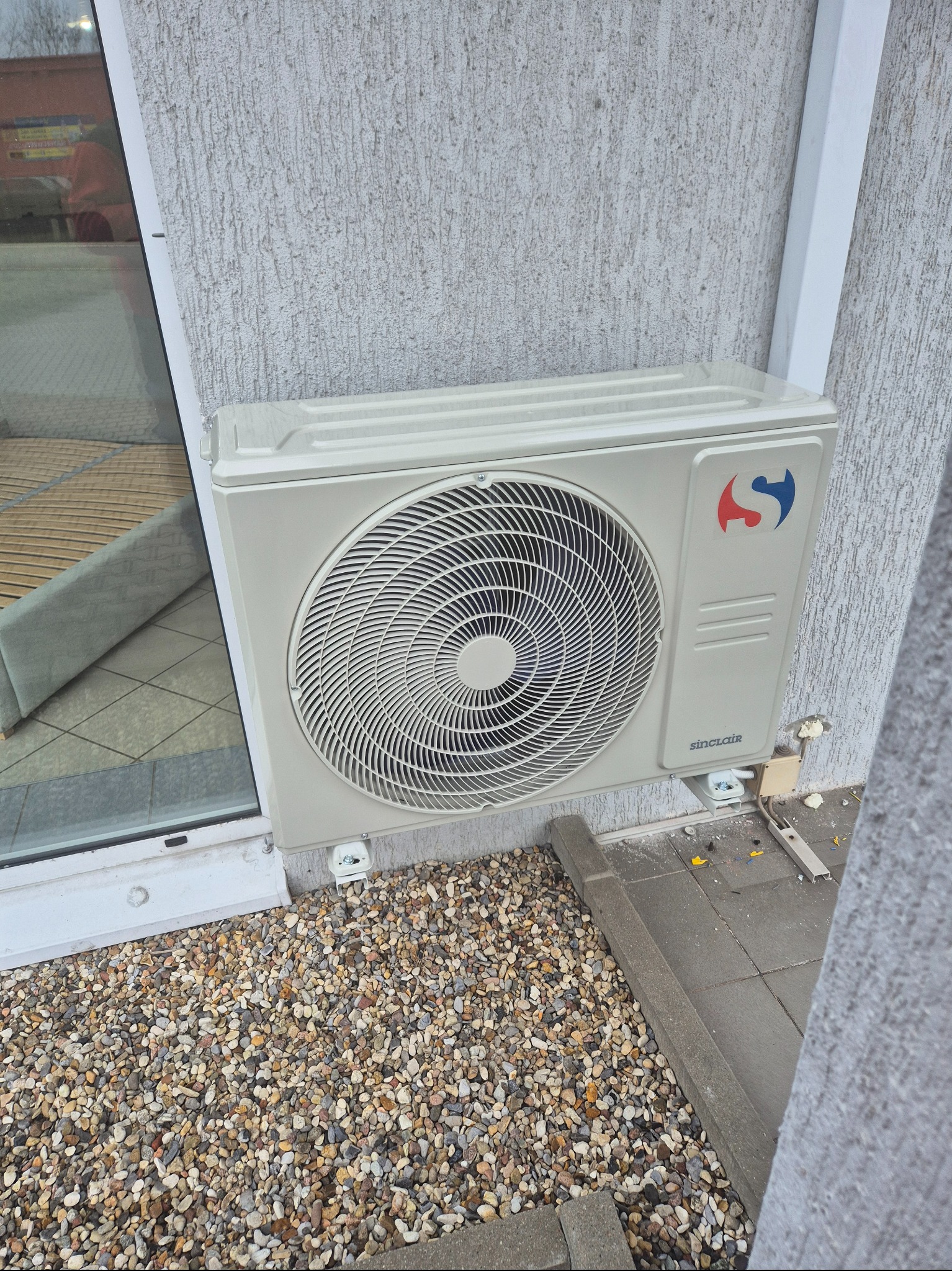 Zamontowana jednostka zewnętrzna klimatyzatora Sinclair na elewacji budynku, widoczne kamyki dekoracyjne i fragment okna z odbiciem wnętrza.