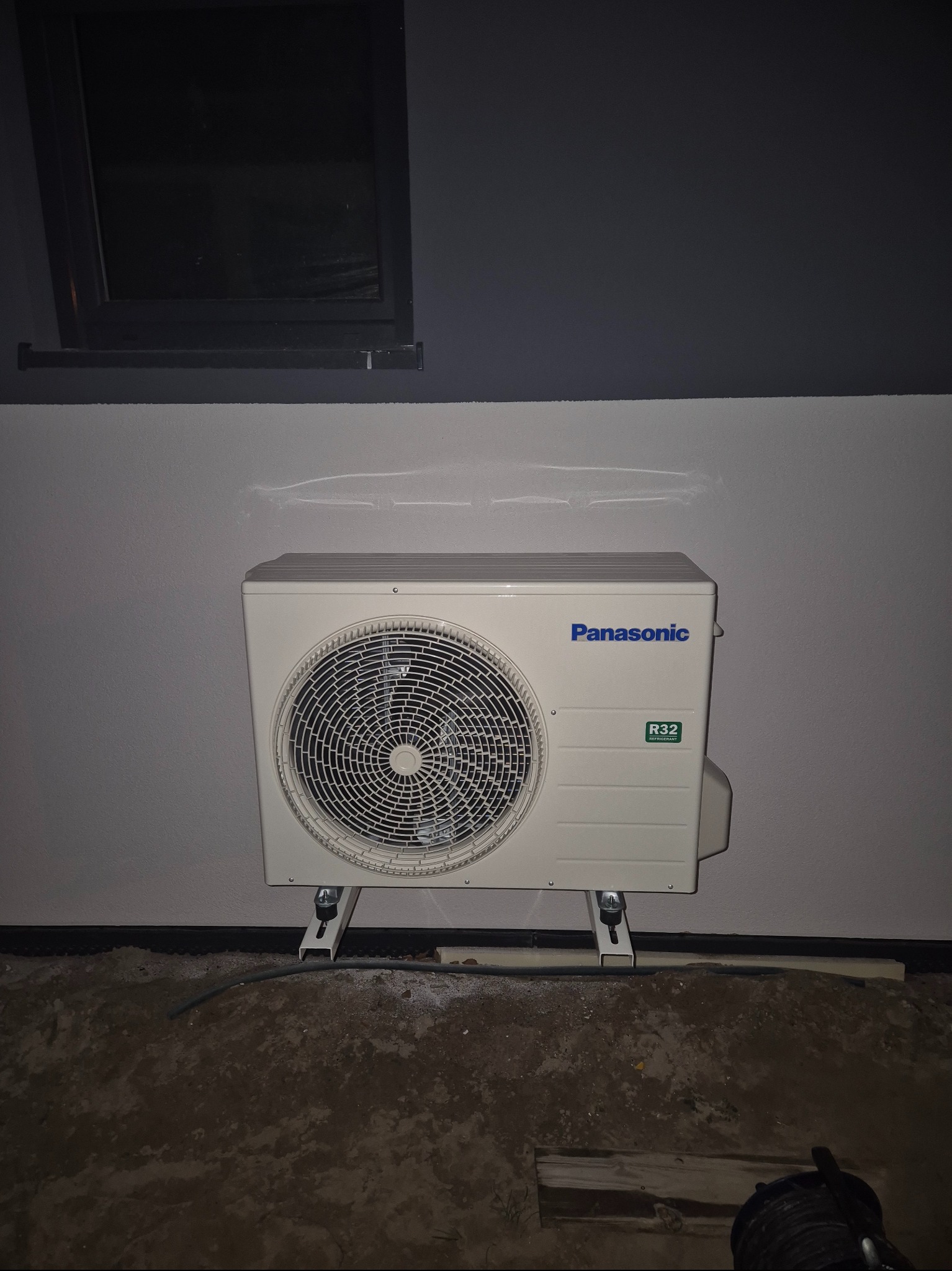 Zewnętrzna jednostka klimatyzacji Panasonic zamontowana na wspornikach przy szarej ścianie, widoczny fragment okna w ciemnej ramie i niedokończona podłoga.