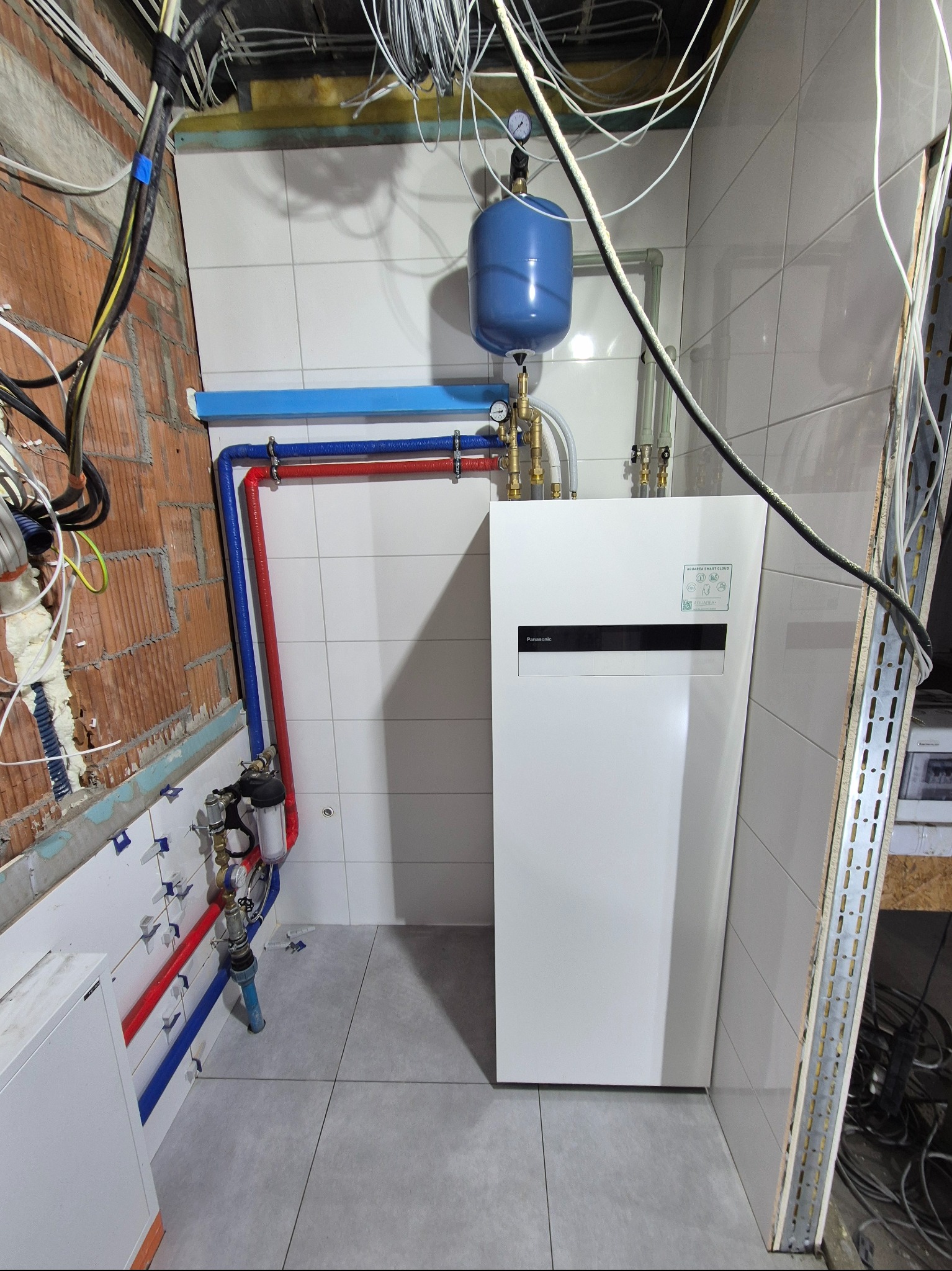 Instalacja pompy ciepła Panasonic Aquarea Smart Cloud w pomieszczeniu z częściowo wykończonymi ścianami, widoczne rury w kolorach czerwonym i niebieskim, zbiornik wyrównawczy oraz przewody...