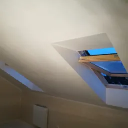 Poddasze w Gadce po nałożeniu gładzi, widoczne dwa okna dachowe z drewnianą ramą i grzejnik na ścianie.