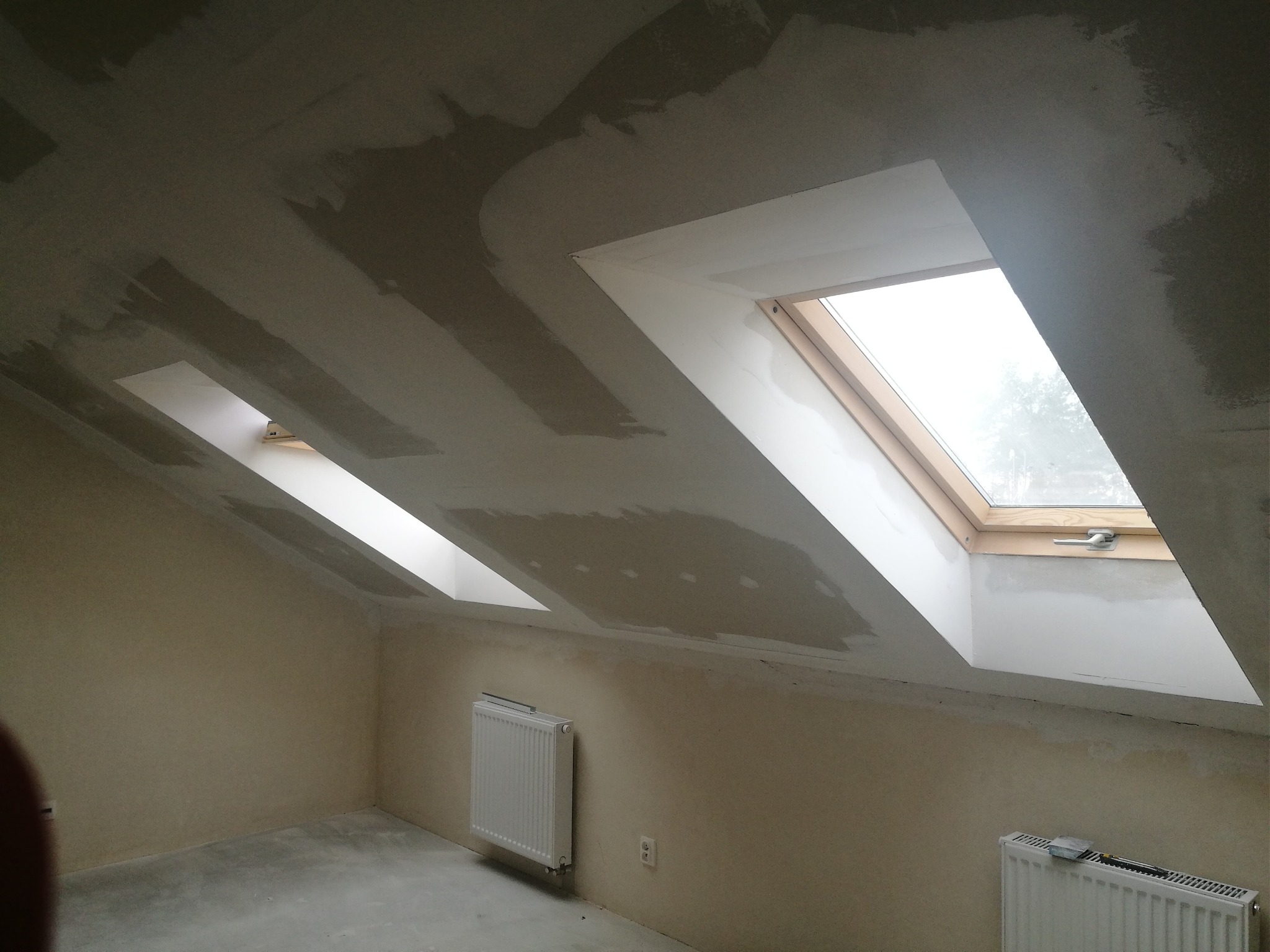 Poddasze w trakcie prac wykończeniowych: ściany i sufit pokryte warstwą gładzi, widoczne dwa okna dachowe i grzejniki.