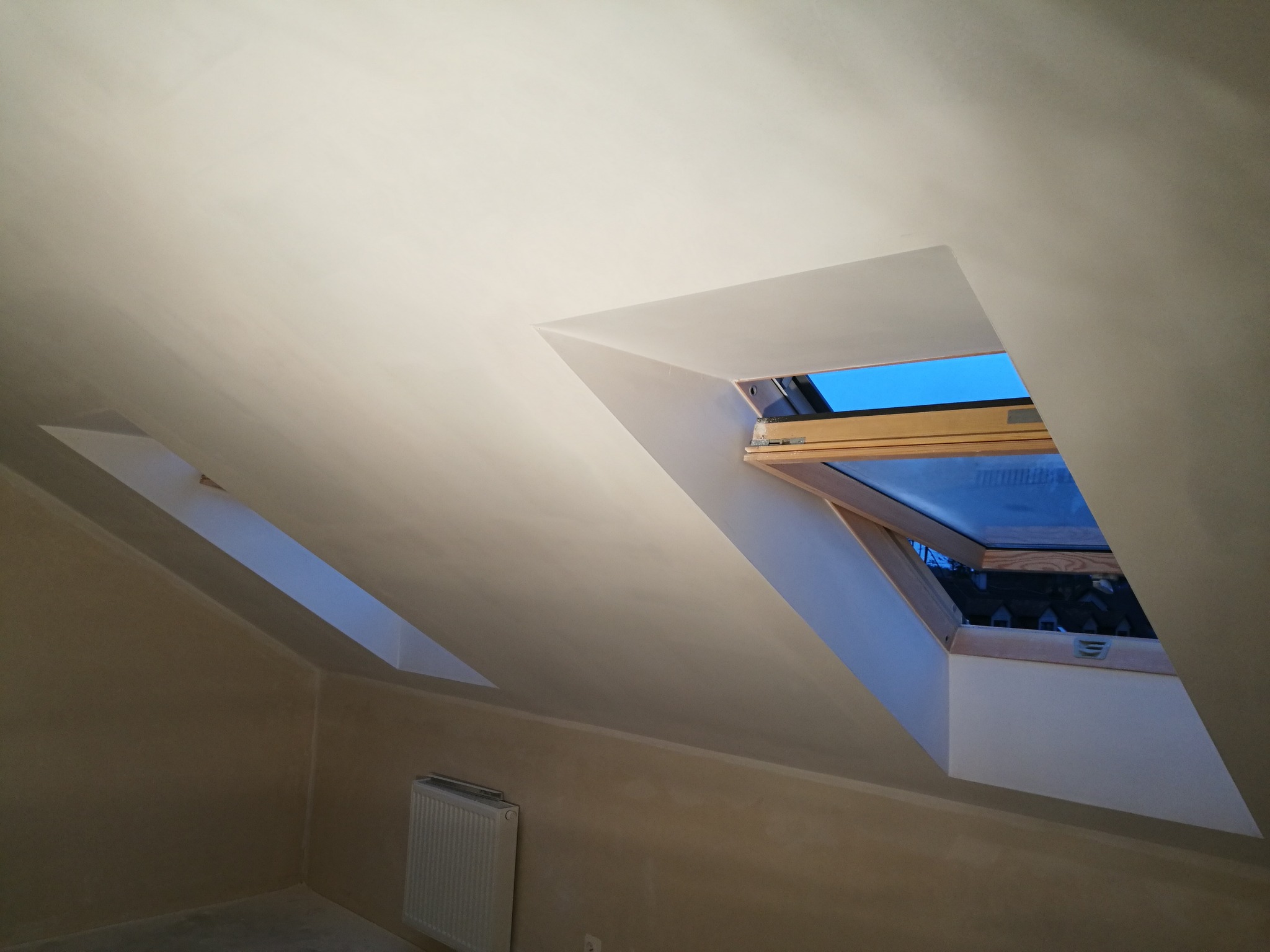 Poddasze w Gadce po nałożeniu gładzi, widoczne dwa okna dachowe z drewnianą ramą i grzejnik na ścianie.