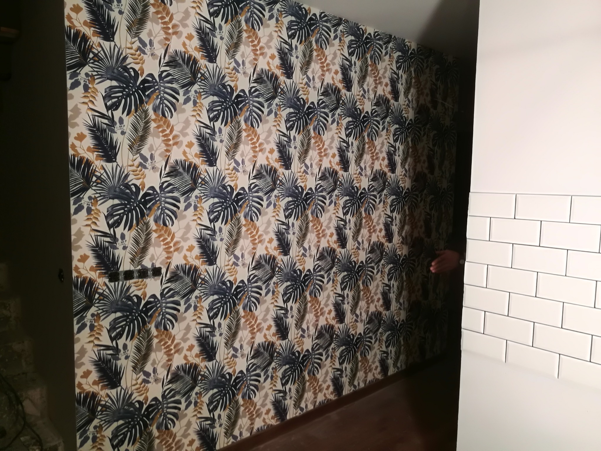 Ściana pokryta tapetą w tropikalne liście, sąsiadująca z fragmentem ściany wyłożonej białymi, prostokątnymi płytkami, widoczny fragment dłoni w przejściu.