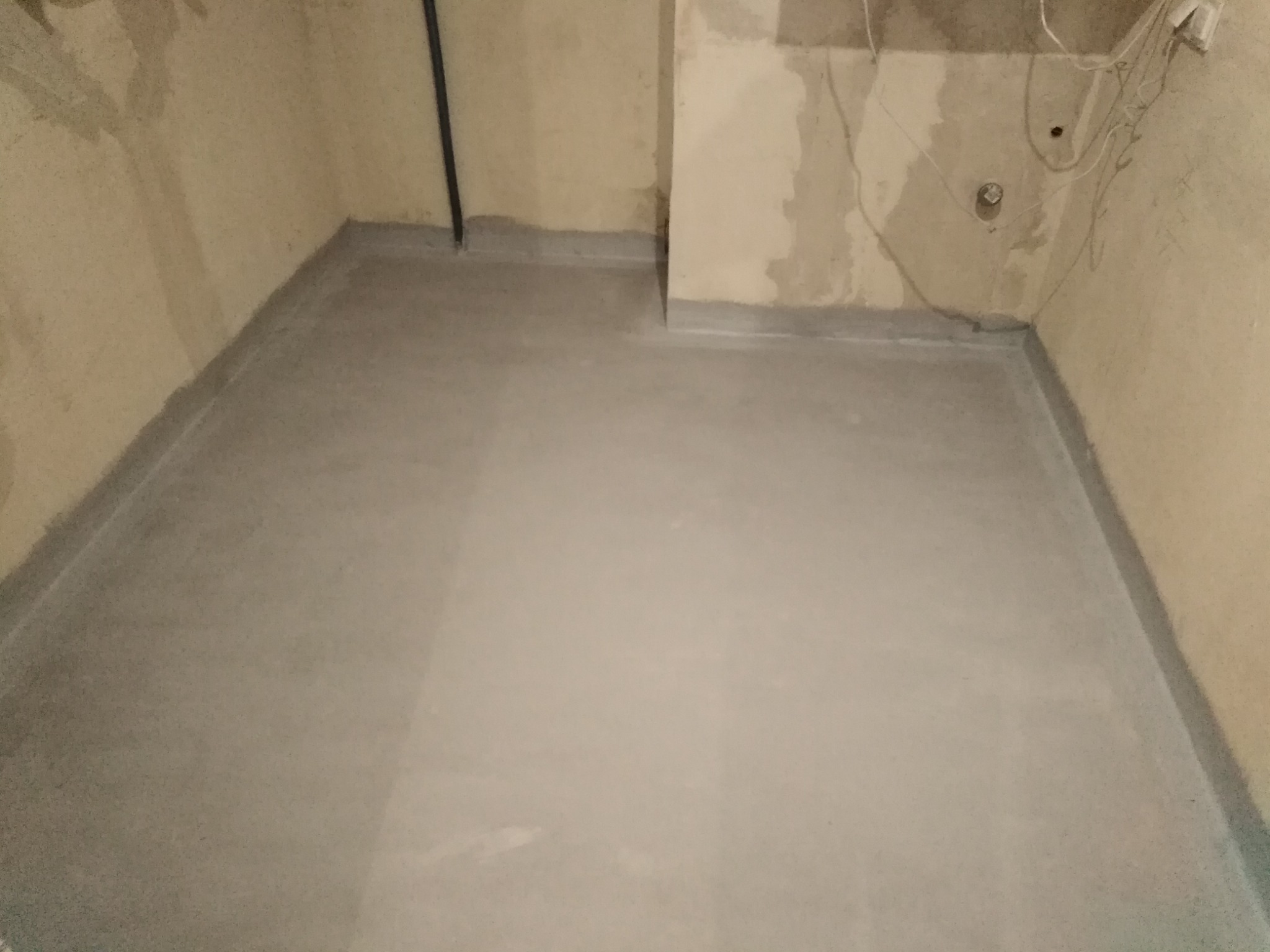 Wyrównana, szara wylewka samopoziomująca na podłodze w pomieszczeniu w trakcie prac remontowych, widoczne niedoskonałości ścian i wystające przewody.