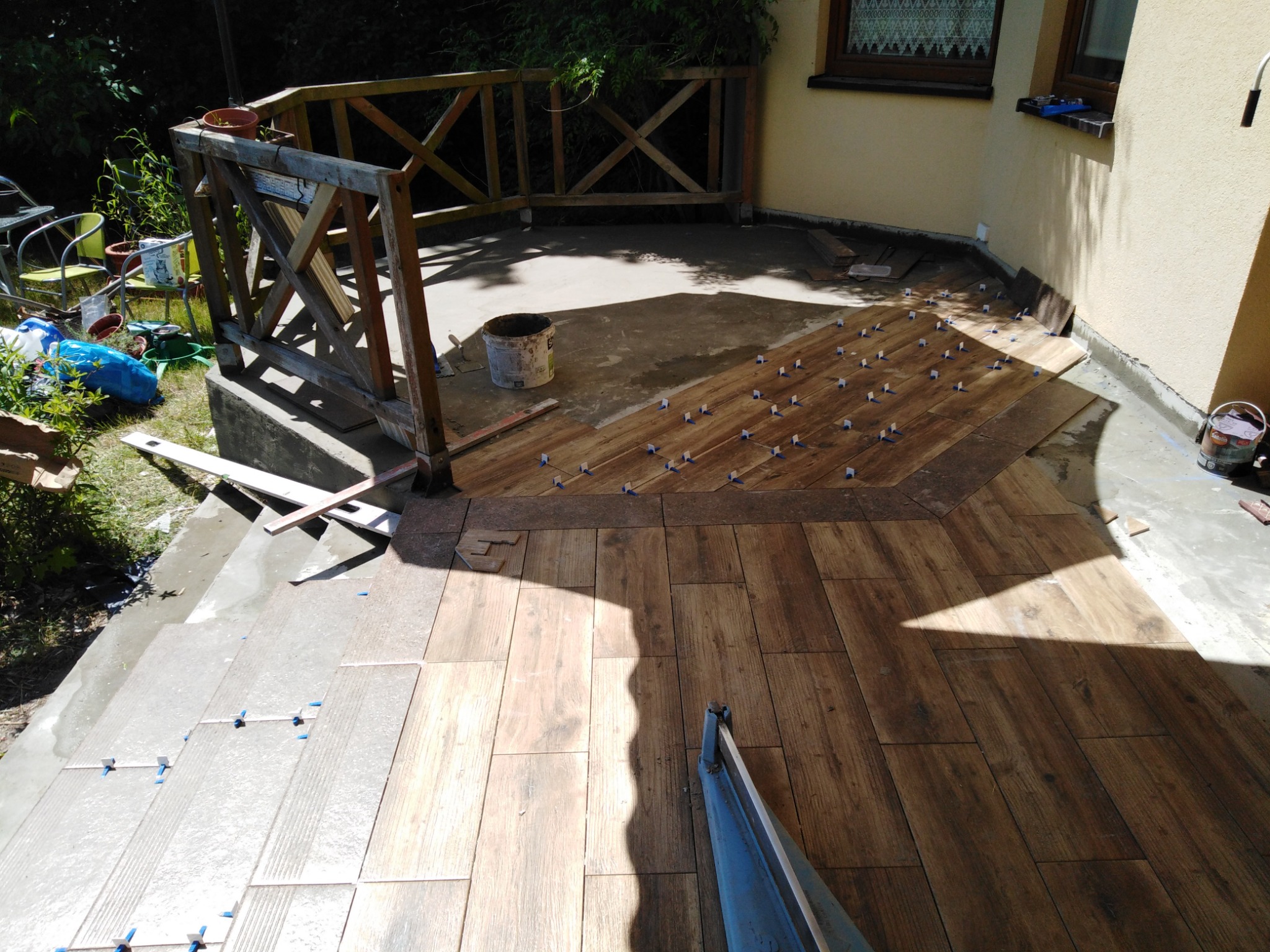 Układanie drewnopodobnych płytek na tarasie, widoczne krzyżyki dystansowe i wiadro z klejem, częściowo ułożona podłoga.
