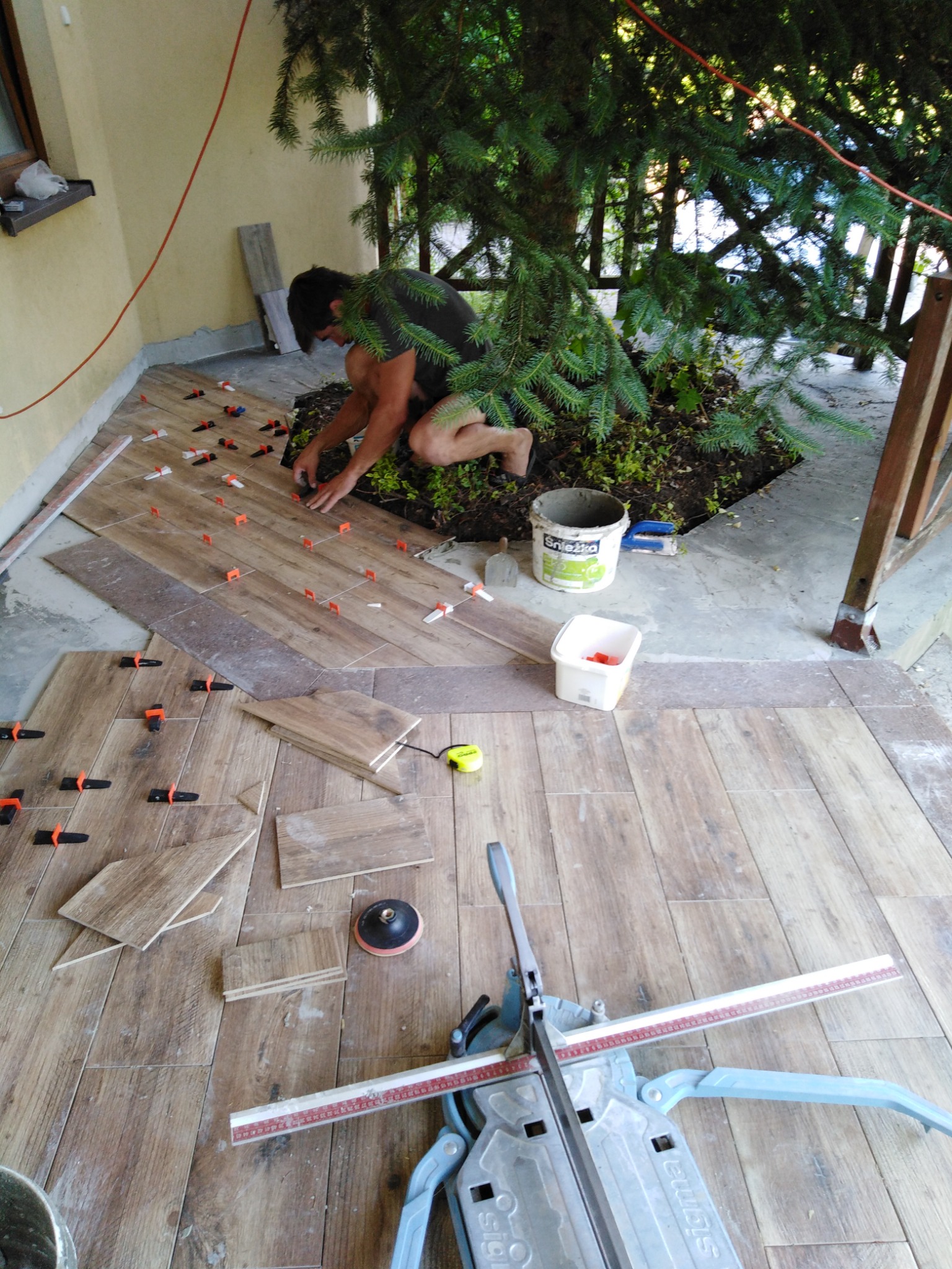 Układanie płytek imitujących drewno na tarasie, widoczny glazurnik przy pracy, poziomice, wiaderko po kleju Śnieżka i narzędzia do cięcia płytek.