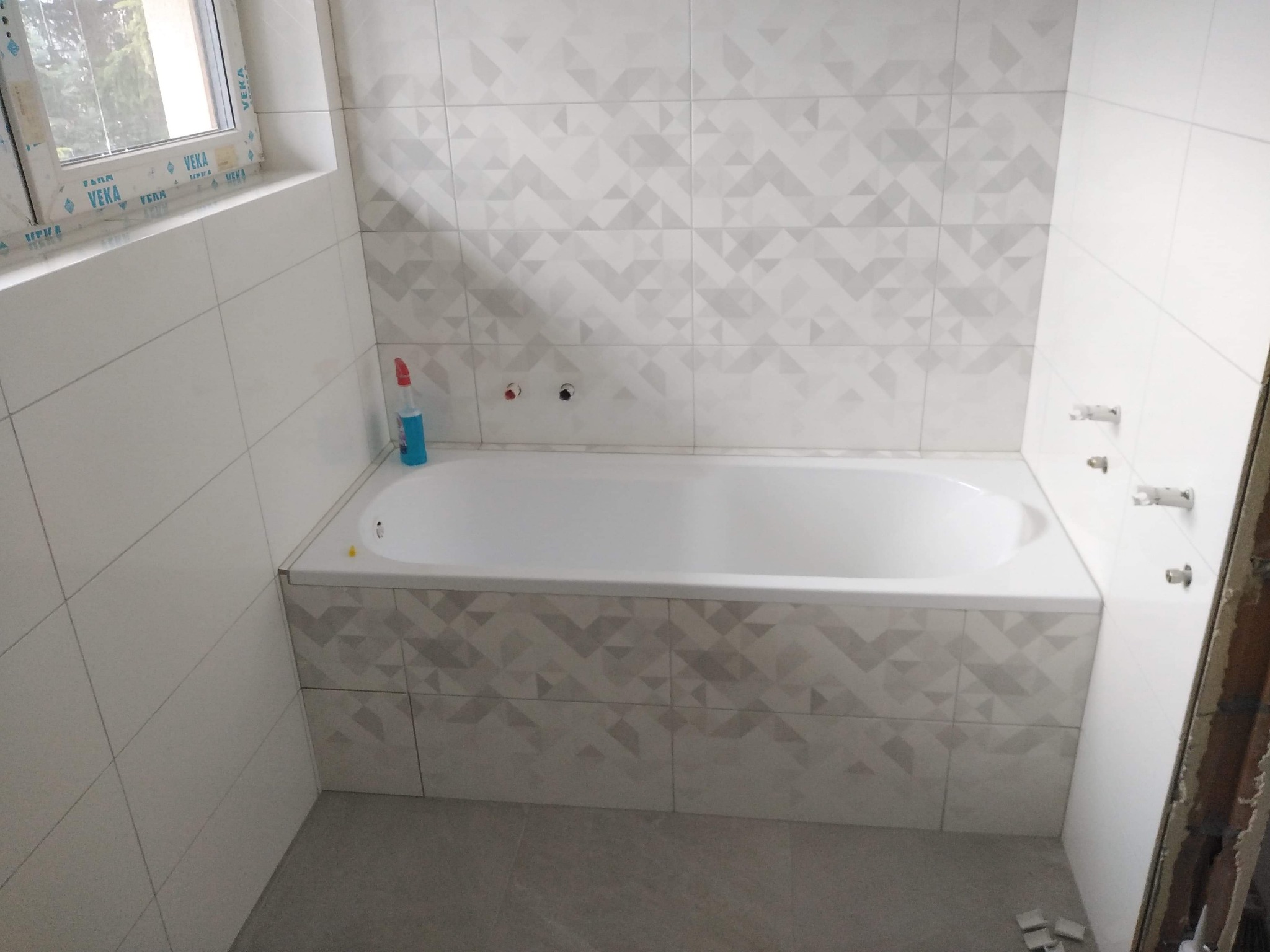 Wanna obudowana płytkami z geometrycznym wzorem, białe płytki na ścianach, okno z widocznym profilem VEKA, butelka płynu do czyszczenia na wannie, szare płytki podłogowe, widoczne przyłącza...