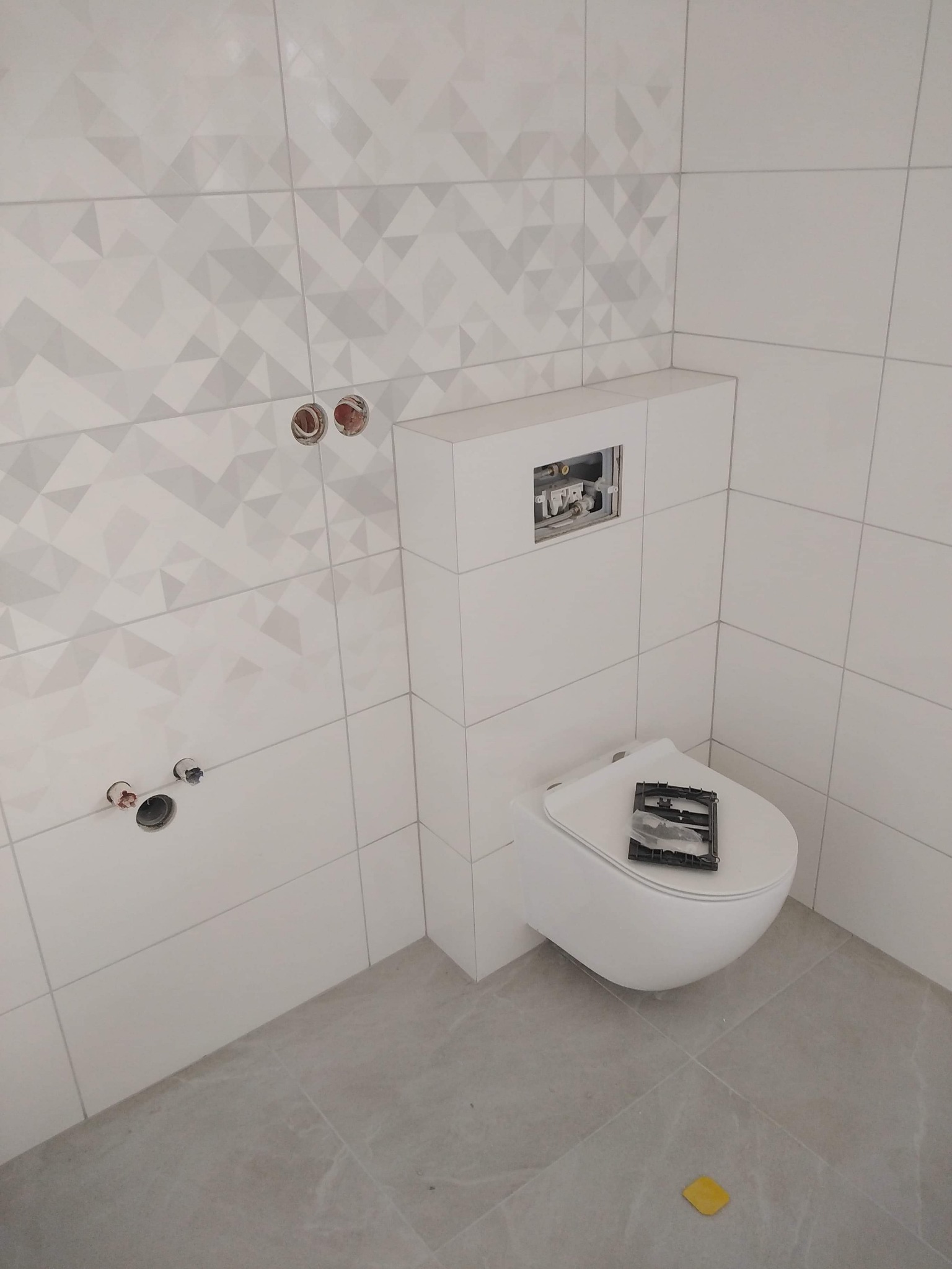 Biała, podwieszana toaleta z elementami montażowymi na desce, na tle ściany z geometrycznymi płytkami i przygotowanymi otworami na armaturę.