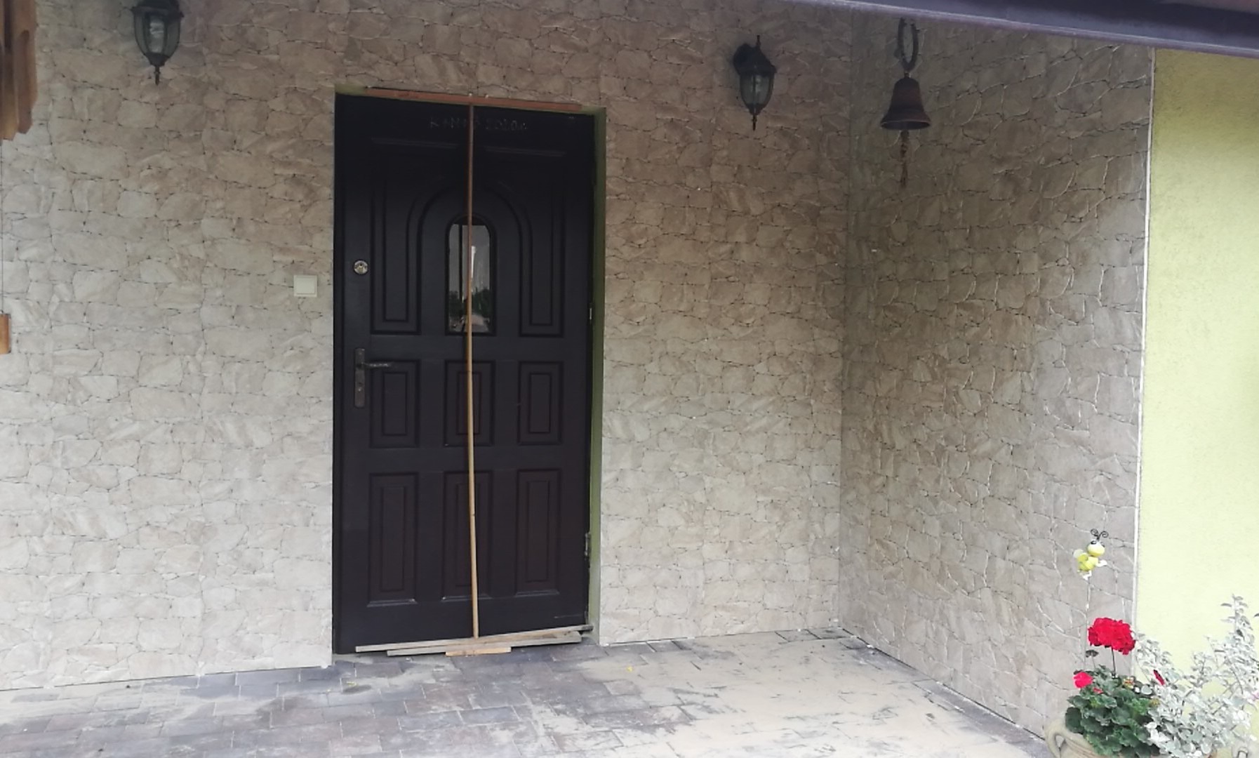 Elewacja zewnętrzna domu wykończona płytkami imitującymi kamień, ciemne drzwi wejściowe zabezpieczone listwami, oświetlenie zewnętrzne w stylu retro, dzwonek i doniczka z czerwonymi kwiatami.