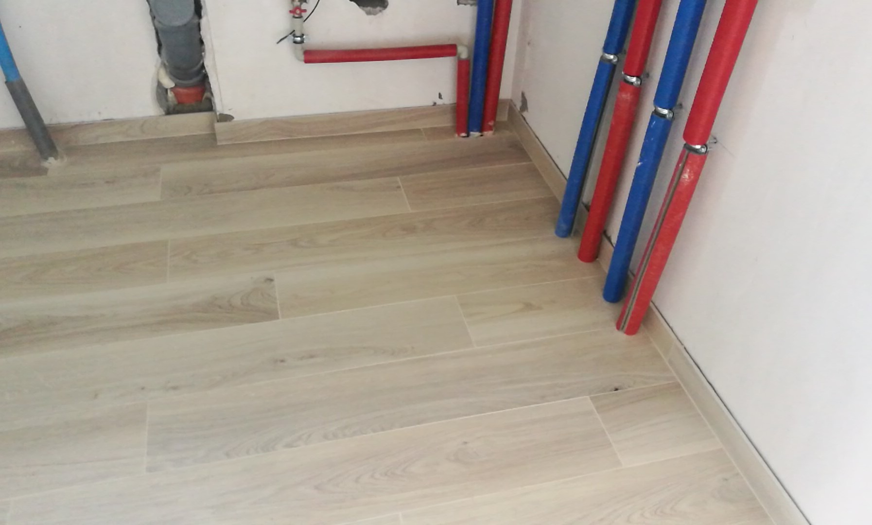 Jasne, podłużne płytki podłogowe imitujące drewno, ułożone w nowej łazience, z widocznymi pionowymi rurami w kolorach czerwonym i niebieskim oraz szarą rurą kanalizacyjną w rogu pomieszczenia.