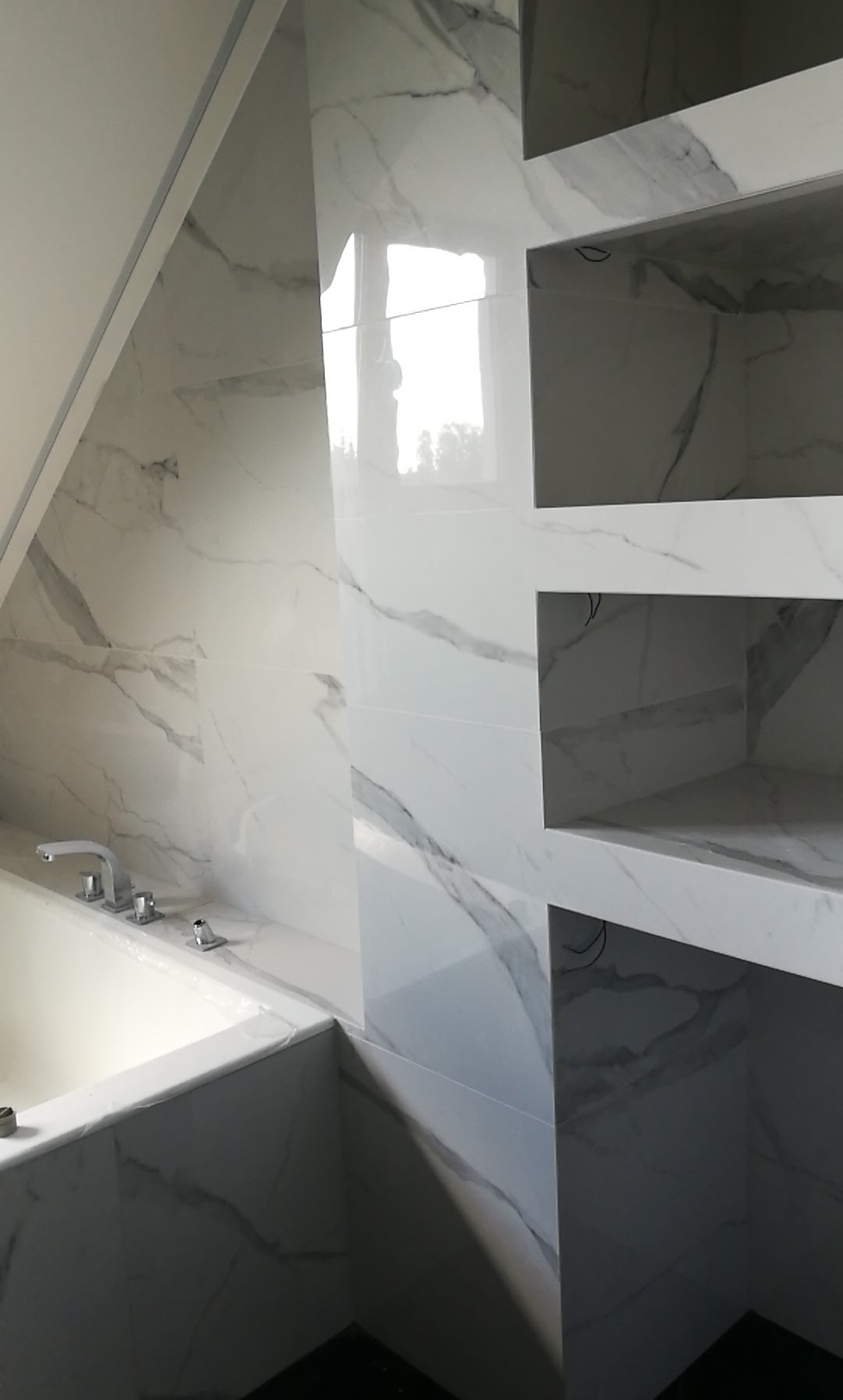 Wykończona łazienka z wanną i półkami wnękowymi wyłożonymi białymi płytkami imitującymi marmur z szarymi żyłami, widoczna bateria wannowa.