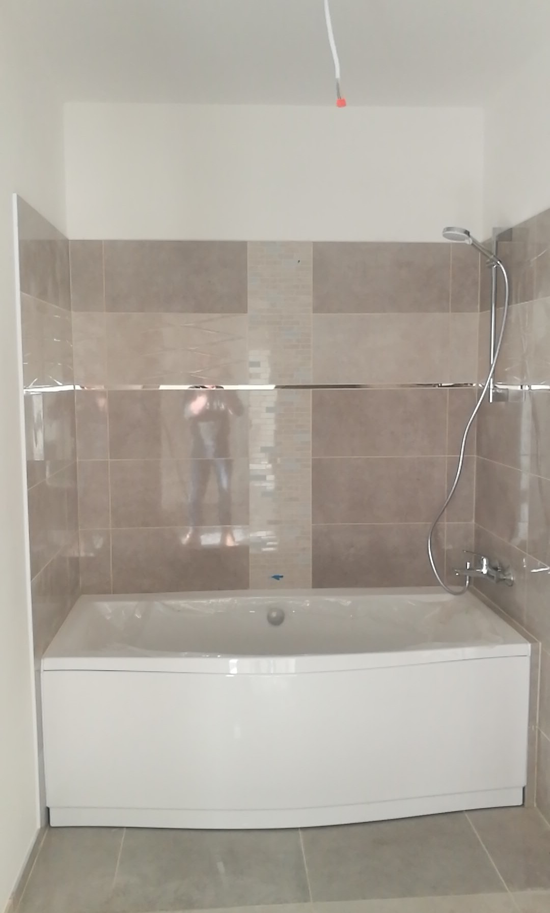 Nowoczesna łazienka z wanną obłożoną beżowymi płytkami, widoczna chromowana armatura i listwa dekoracyjna, odblask osoby fotografującej w lustrzanej powierzchni.