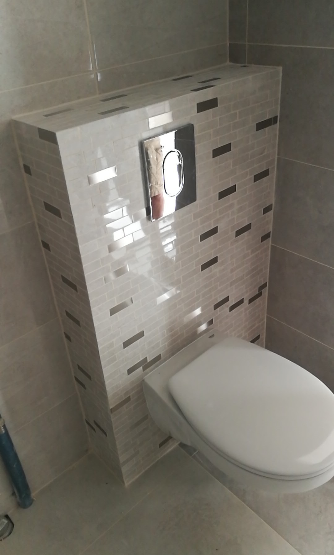 Podwieszana toaleta z zabudową stelaża pokrytą jasnoszarą mozaiką z poziomymi, ciemniejszymi akcentami. Widoczny chromowany przycisk spłukiwania i szare płytki podłogowe.