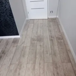 Ułożone panele podłogowe imitujące jasne drewno, widoczne przy ścianie z czarną wzorzystą tapetą i białą listwą przypodłogową oraz białe drzwi w tle.