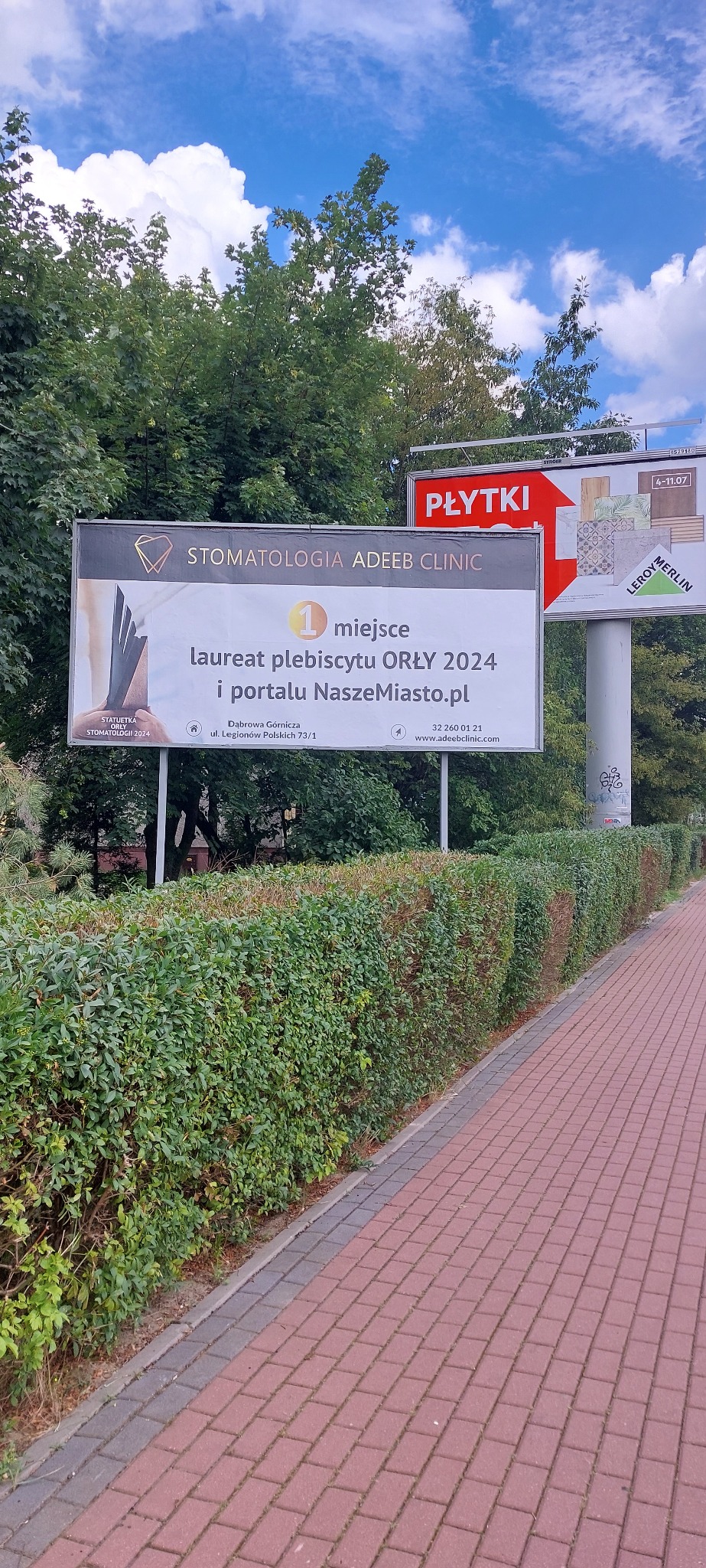 Billboard reklamowy kliniki stomatologicznej Adeeb Clinic, laureata plebiscytu Orły 2024, zlokalizowany przy ulicy Legionów Polskich w Dąbrowie Górniczej, widoczny z chodnika.
