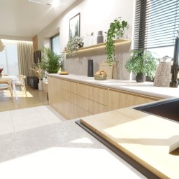 Kliminowska Design - Jasna kuchnia z drewnianymi szafkami, blatem z szarego kamienia i roślinami doniczkowymi na półce. W tle widoczny stół z krzesłami i zasłonięte okno.