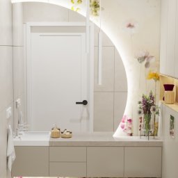 Kliminowska Design - Nowoczesna łazienka z podświetlanym lustrem w kształcie półksiężyca, kwiatowym motywem na ścianie i beżową szafką pod umywalką z podświetleniem LED. Widoczny biały taboret i wisząca toaleta.