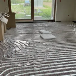 Białe rurki ogrzewania podłogowego ułożone na czerwonej siatce, widoczne dwa betonowe bloki i okno z widokiem na zewnątrz.