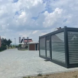 Szara, nowoczesna konstrukcja z przeszkleniami i poziomymi żaluzjami na tle brukowanej nawierzchni, z widocznymi budynkami w oddali i zachmurzonym niebem.