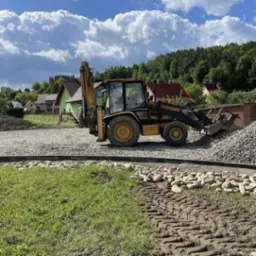 Żółto-czarna koparko-ładowarka na placu budowy z wysypanym żwirem, w tle zabudowa jednorodzinna i zalesione wzgórze.