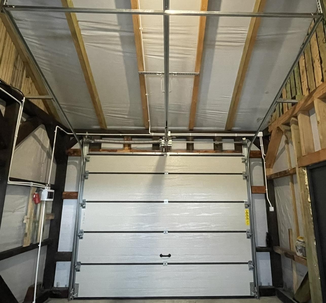 Zamknięta, segmentowa brama garażowa w jasnym kolorze, widoczna od wewnątrz, z widocznymi elementami konstrukcyjnymi garażu i instalacją elektryczną.