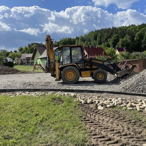 Żółto-czarna koparko-ładowarka na placu budowy z wysypanym żwirem, w tle zabudowa jednorodzinna i zalesione wzgórze.
