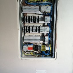 Elektryk Rokietnica - ELEKTRO-PROGRES - Instalacje Elektryczne - Rozdzielnica elektryczna wnękowa z zainstalowanymi bezpiecznikami, przekaźnikami i okablowaniem w trakcie montażu, widoczny symbol uziemienia na ścianie.