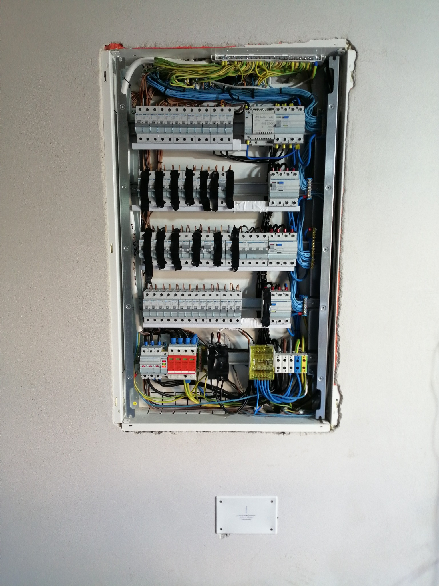 Rozdzielnica elektryczna wnękowa z zainstalowanymi bezpiecznikami, przekaźnikami i okablowaniem w trakcie montażu, widoczny symbol uziemienia na ścianie.
