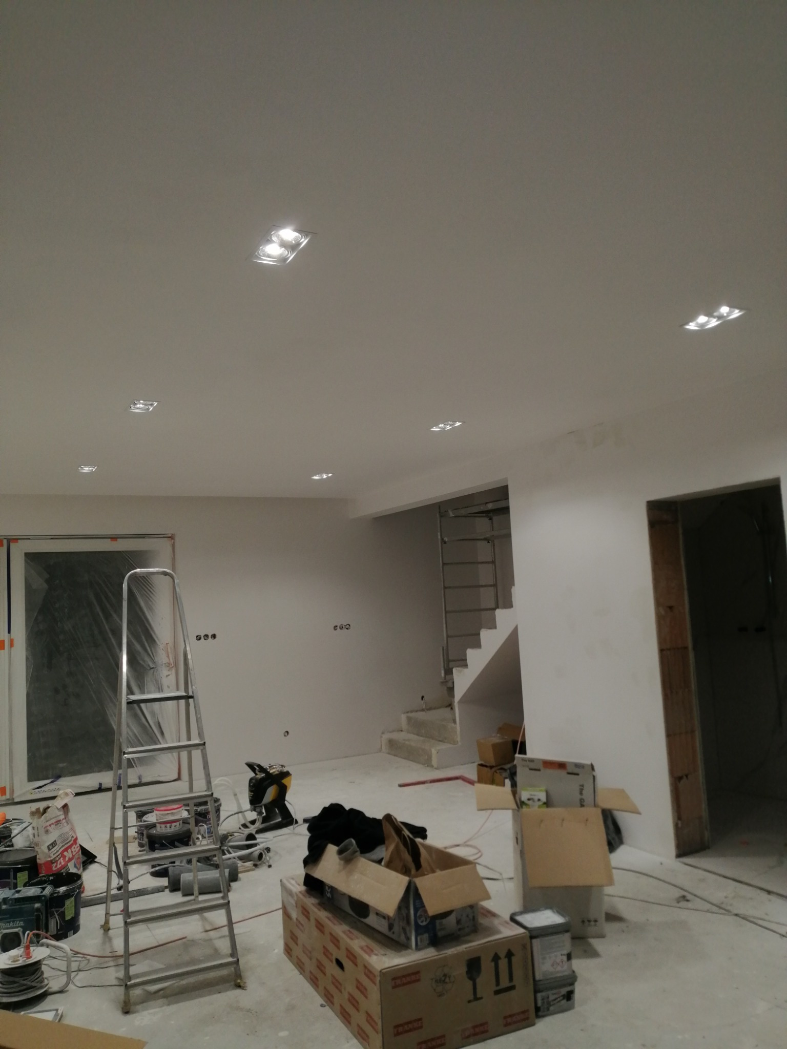 Wnętrze w trakcie remontu z zamontowanymi punktowymi oprawami oświetleniowymi w suficie, widoczne narzędzia, drabina oraz materiały budowlane w kartonach.