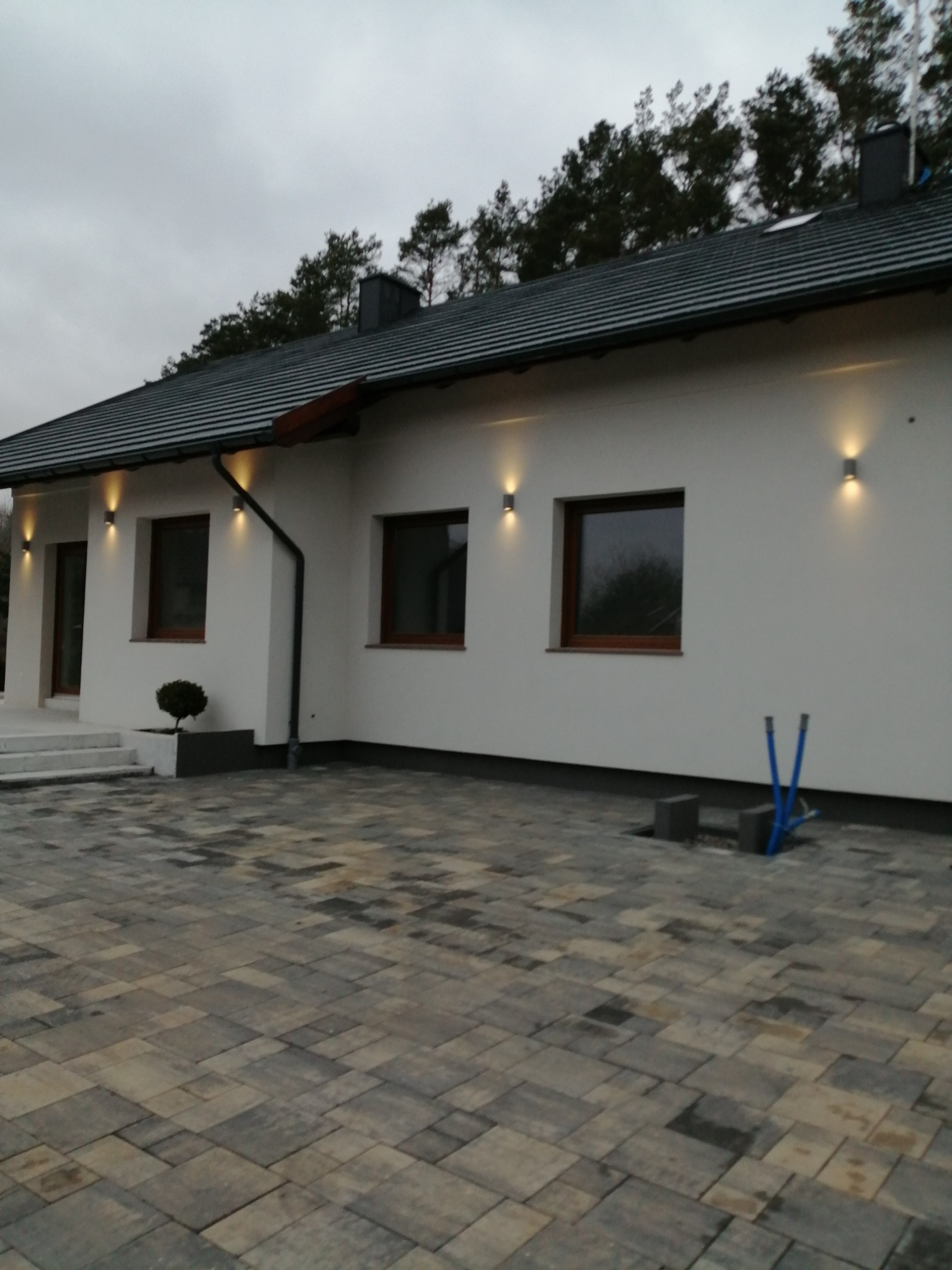 Nowoczesny dom jednorodzinny z oświetleniem elewacyjnym, widoczne okna z brązowymi ramami, kostka brukowa na podjeździe i elementy instalacji elektrycznej na zewnątrz.