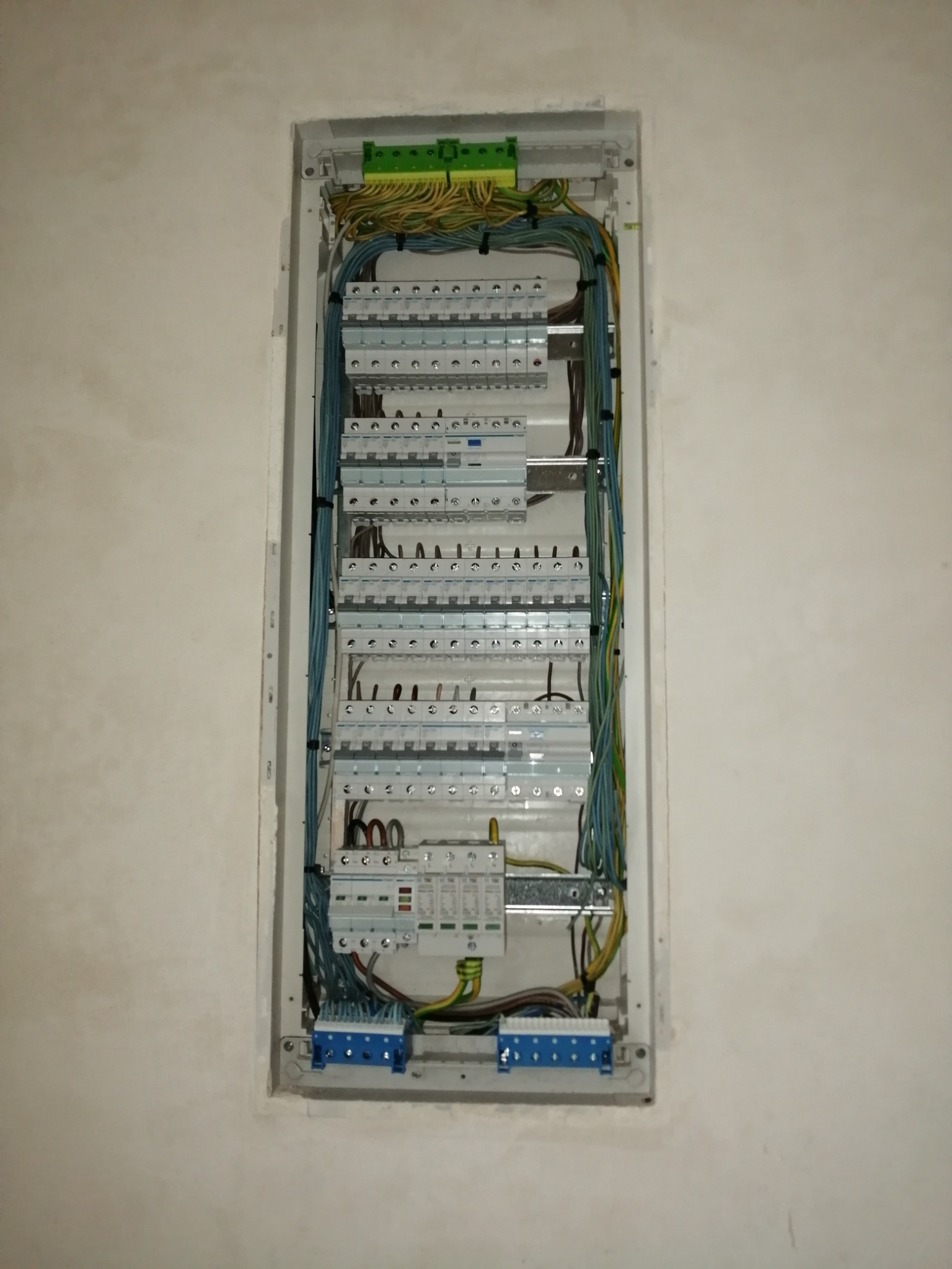 Rozdzielnica elektryczna z widocznymi bezpiecznikami, przewodami w różnych kolorach i listwami zaciskowymi, zamontowana na jasnej ścianie.