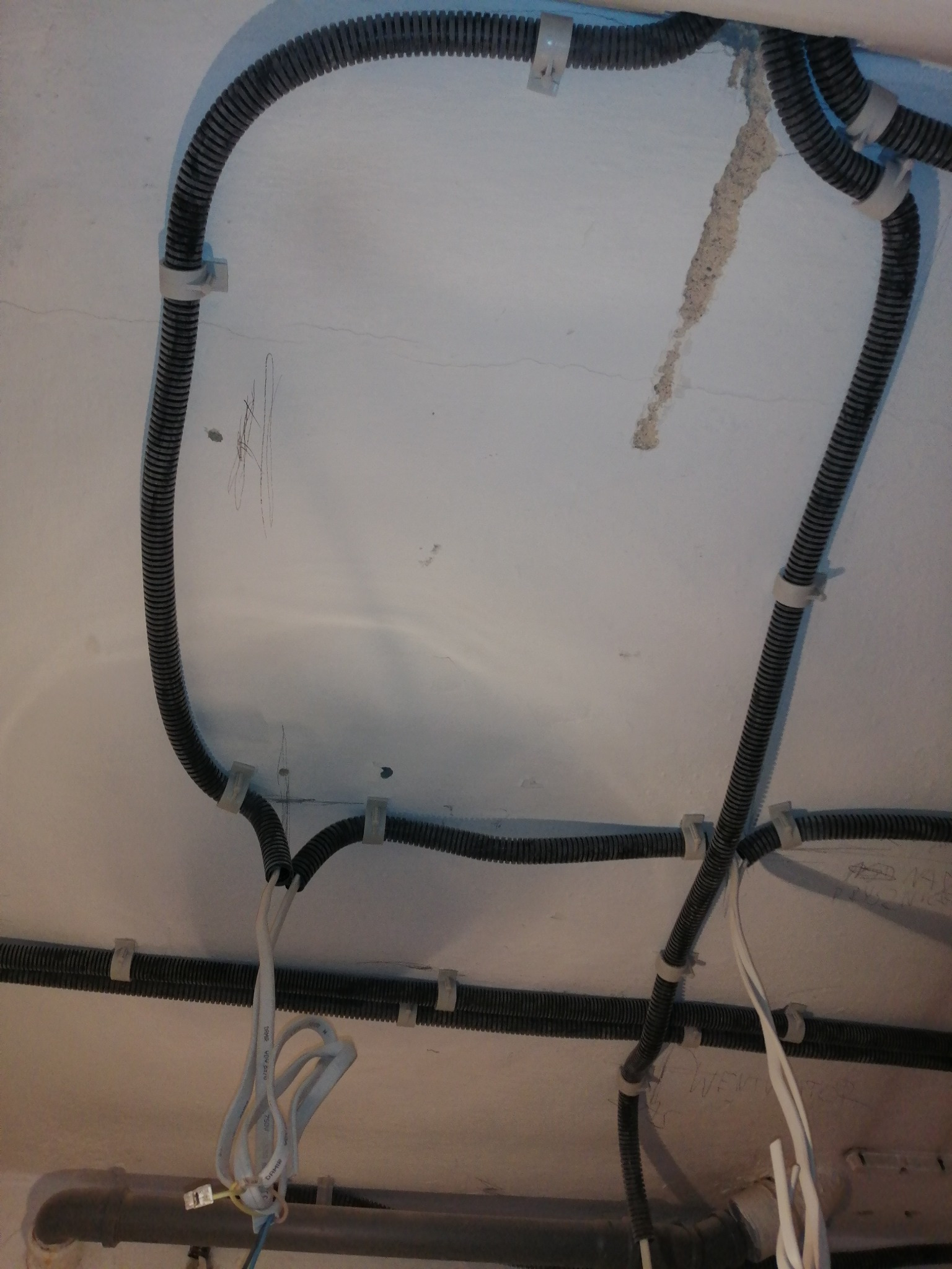 Kable elektryczne w czarnej karbowanej rurze, przymocowane białymi uchwytami do sufitu z widocznymi śladami po instalacjach i zabrudzeniami.