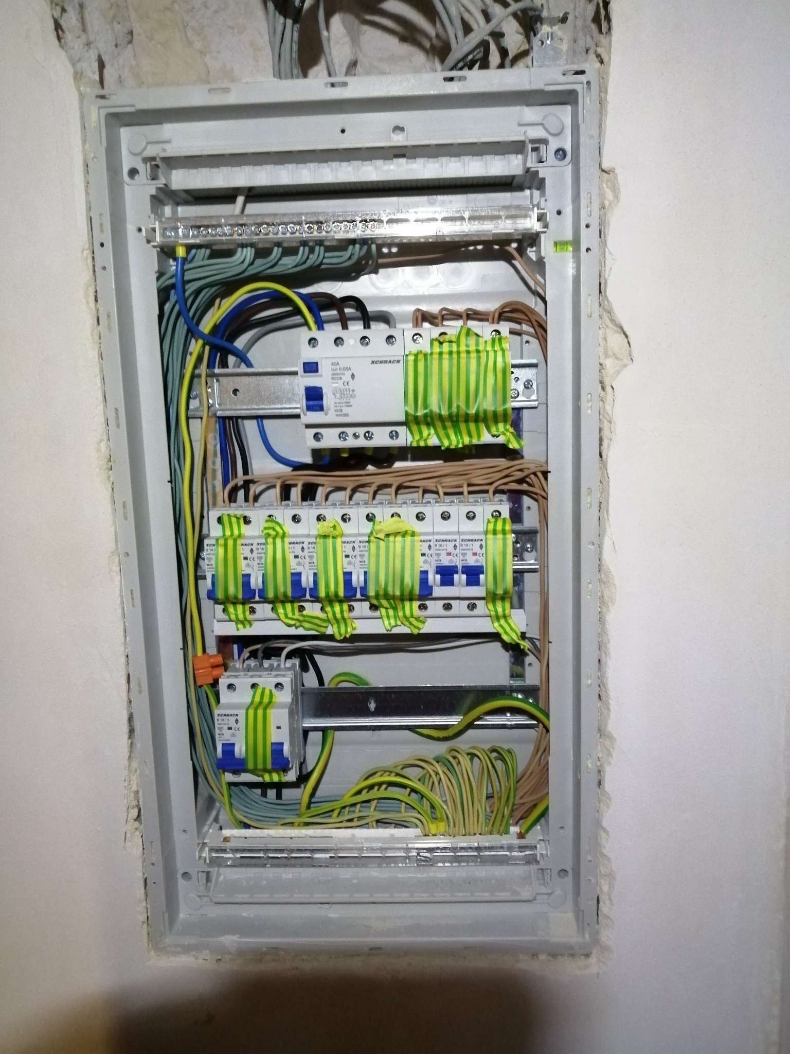Rozdzielnica elektryczna z zamontowanymi bezpiecznikami, przewodami w różnych kolorach, osadzona w ścianie z widocznymi śladami po montażu.