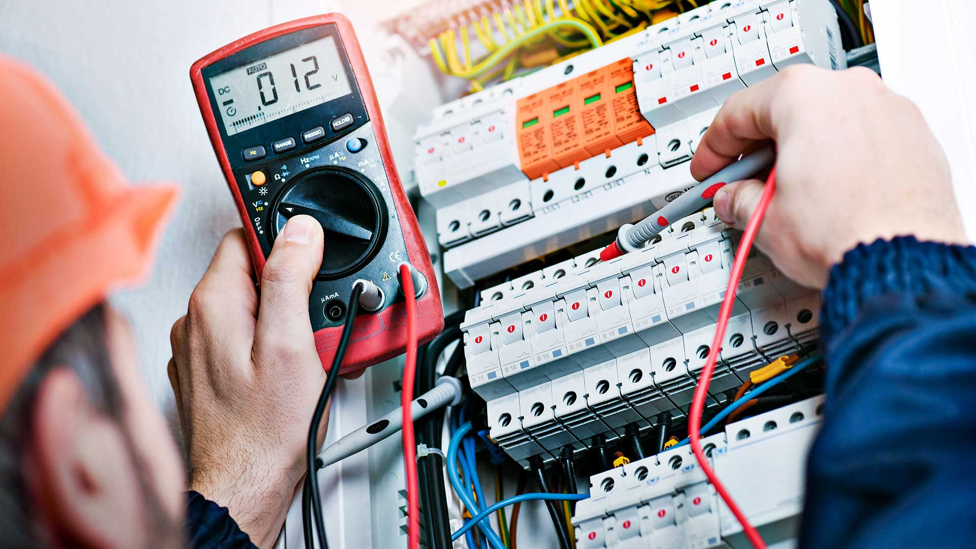 Elektryk w kasku mierzy napięcie w rozdzielnicy za pomocą multimetru cyfrowego, widoczne przewody i bezpieczniki.