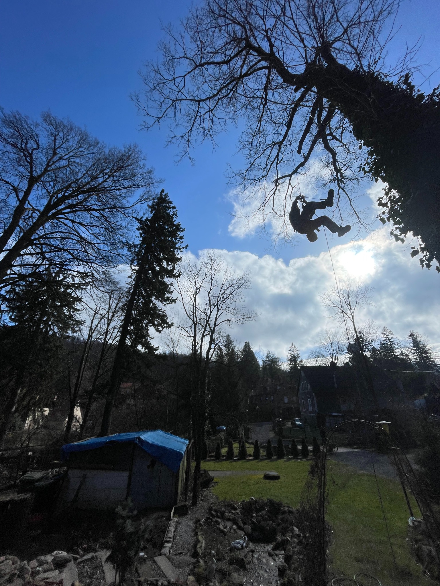 Sylwetka pracownika na linie, zawieszona na gałęzi drzewa na tle błękitnego nieba z chmurami, widok z dołu na teren z zadaszoną konstrukcją i zielonym trawnikiem.