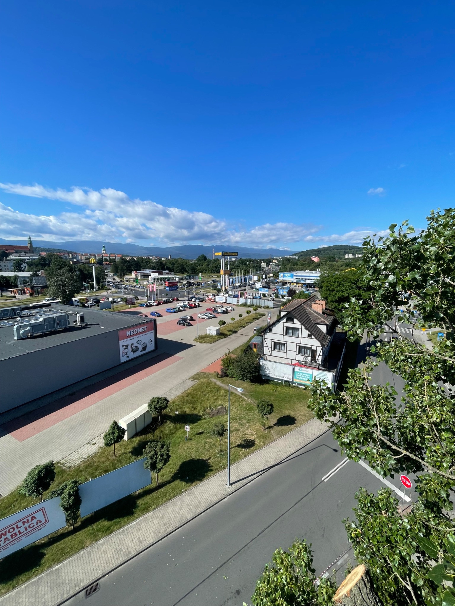 Widok z góry na miasto z widocznym parkingiem, budynkami usługowymi i drzewami po prawej stronie, w tym pień po świeżo ściętym drzewie, na tle błękitnego nieba z chmurami.
