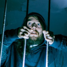Fotografiedudy - Mężczyzna z brodą i w kapturze, z przerażeniem w oczach, trzyma się krat zza potłuczonego szkła, oświetlony od dołu niebieskim światłem.