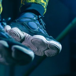 Fotografiedudy - Zbliżenie na podeszwę i fragment cholewki sportowego buta w odcieniach granatu i bieli, z widoczną teksturą podeszwy przypominającą labirynt, oraz fragment spodni w żółto-czarny wzór na górze kadru.