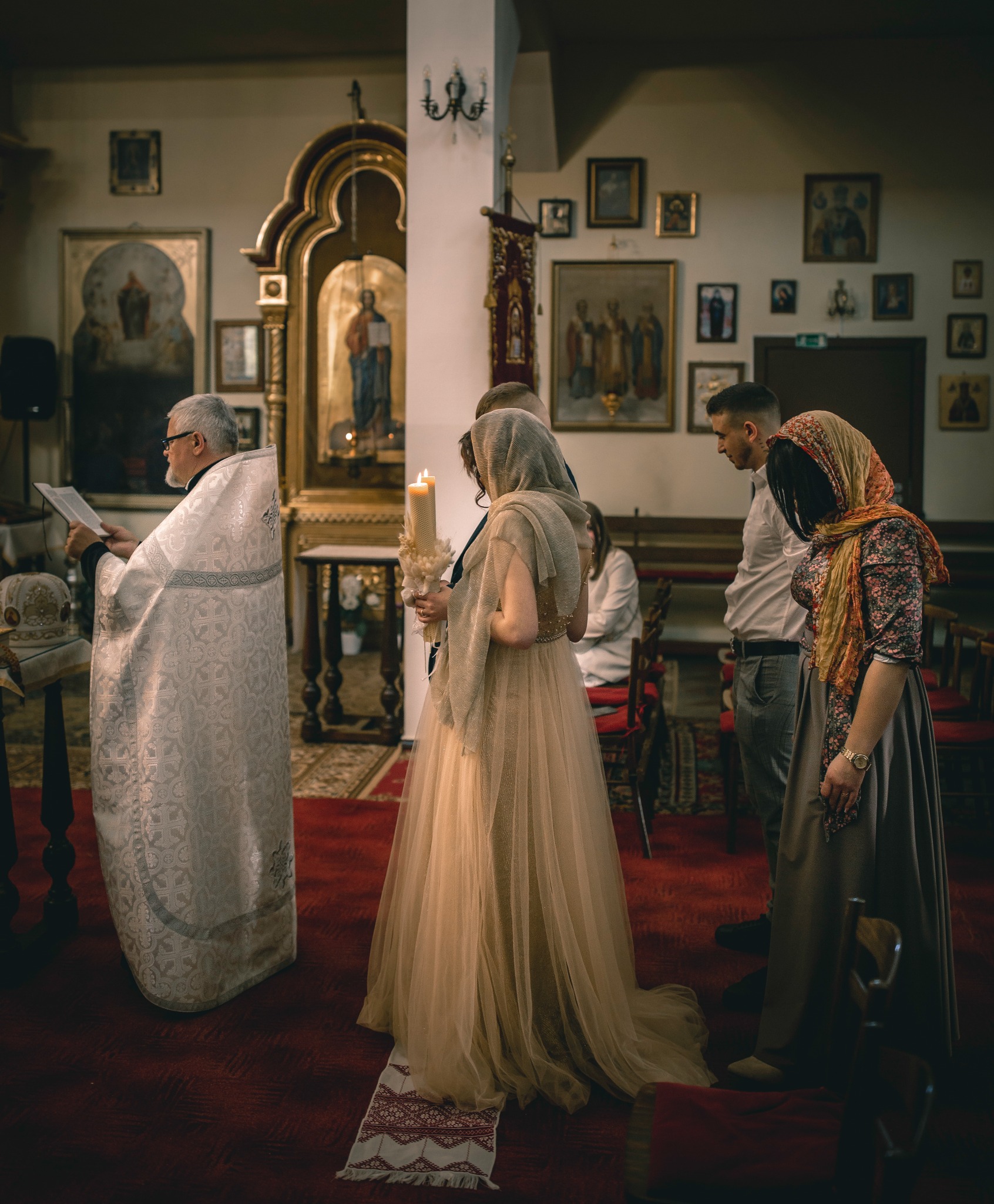 Uroczystość w cerkwi: kobieta w sukni i welonie trzyma zapaloną świecę, obok ksiądz czyta z księgi, w tle ikony i obrazy.