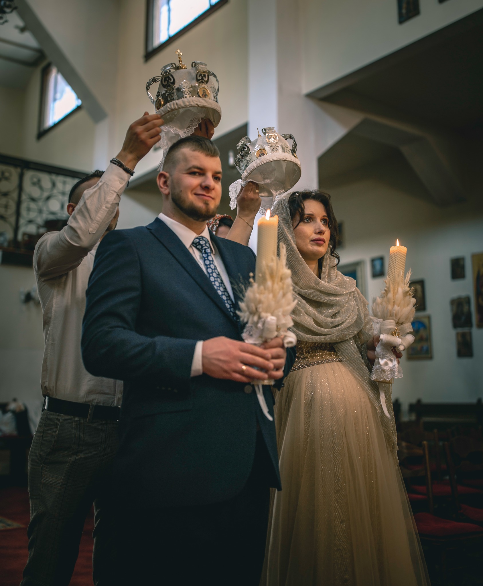 Para młoda podczas ceremonii zaślubin w kościele, z koronami trzymanymi nad głowami i świecami w dłoniach.