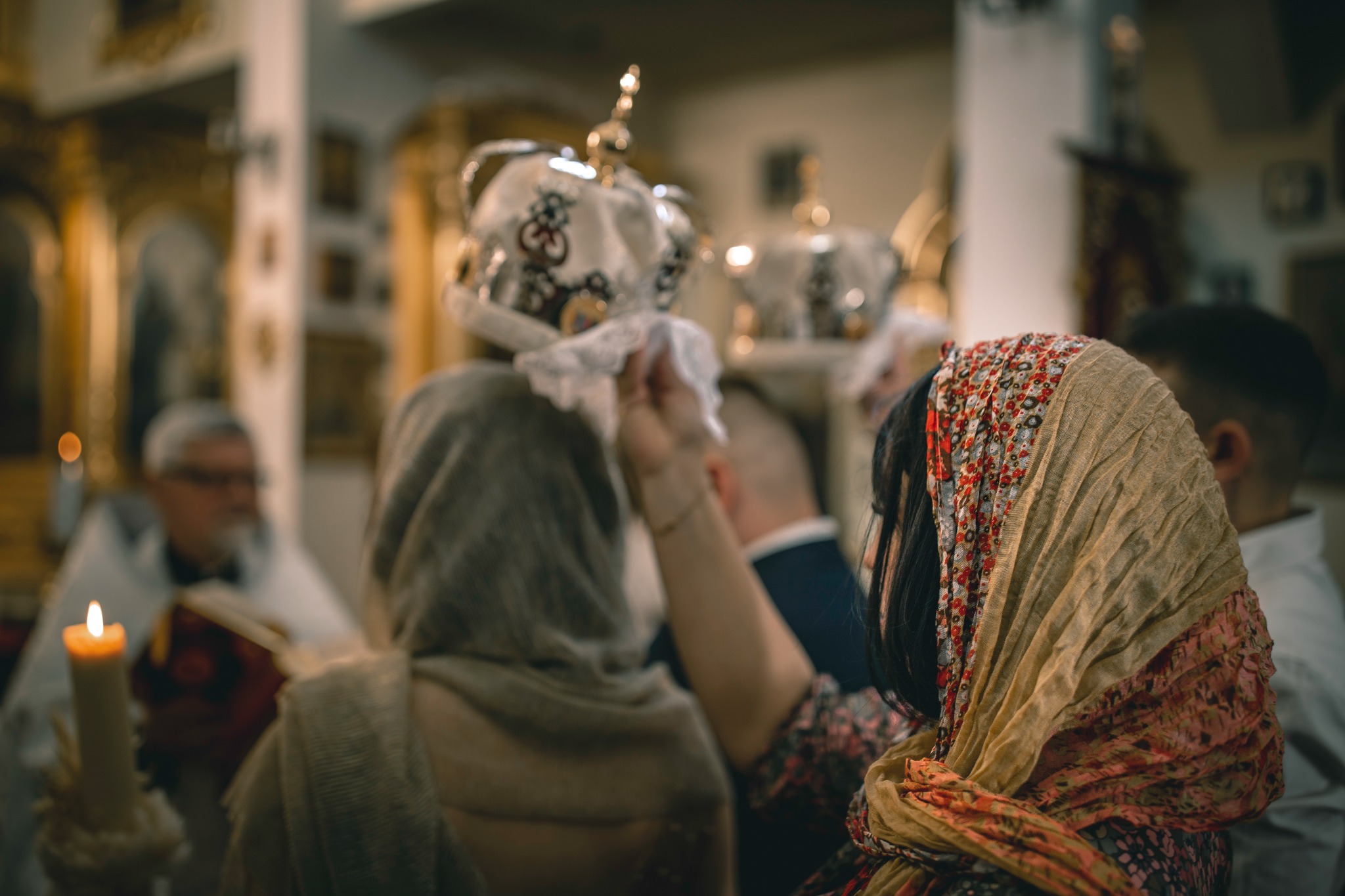 Wnętrze cerkwi podczas ceremonii ślubnej, widoczne korony ślubne na głowach pary młodej, ksiądz z płonącą świecą w tle.
