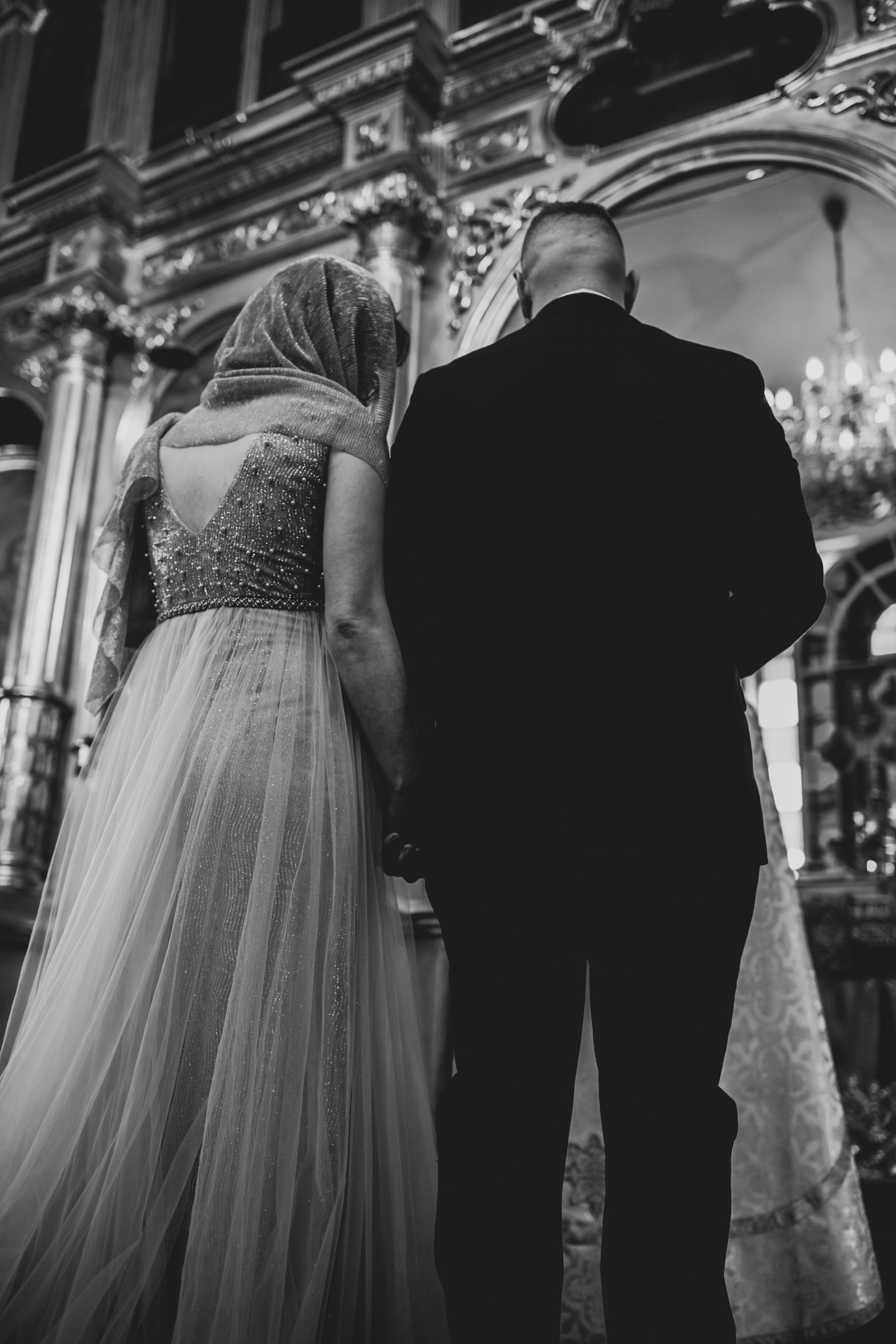 Para młoda trzymająca się za ręce w kościele, widok z tyłu, ona w sukni z odkrytymi plecami i szalu, on w czarnym garniturze, bogato zdobione wnętrze kościoła w tle, czarno-białe zdjęcie.