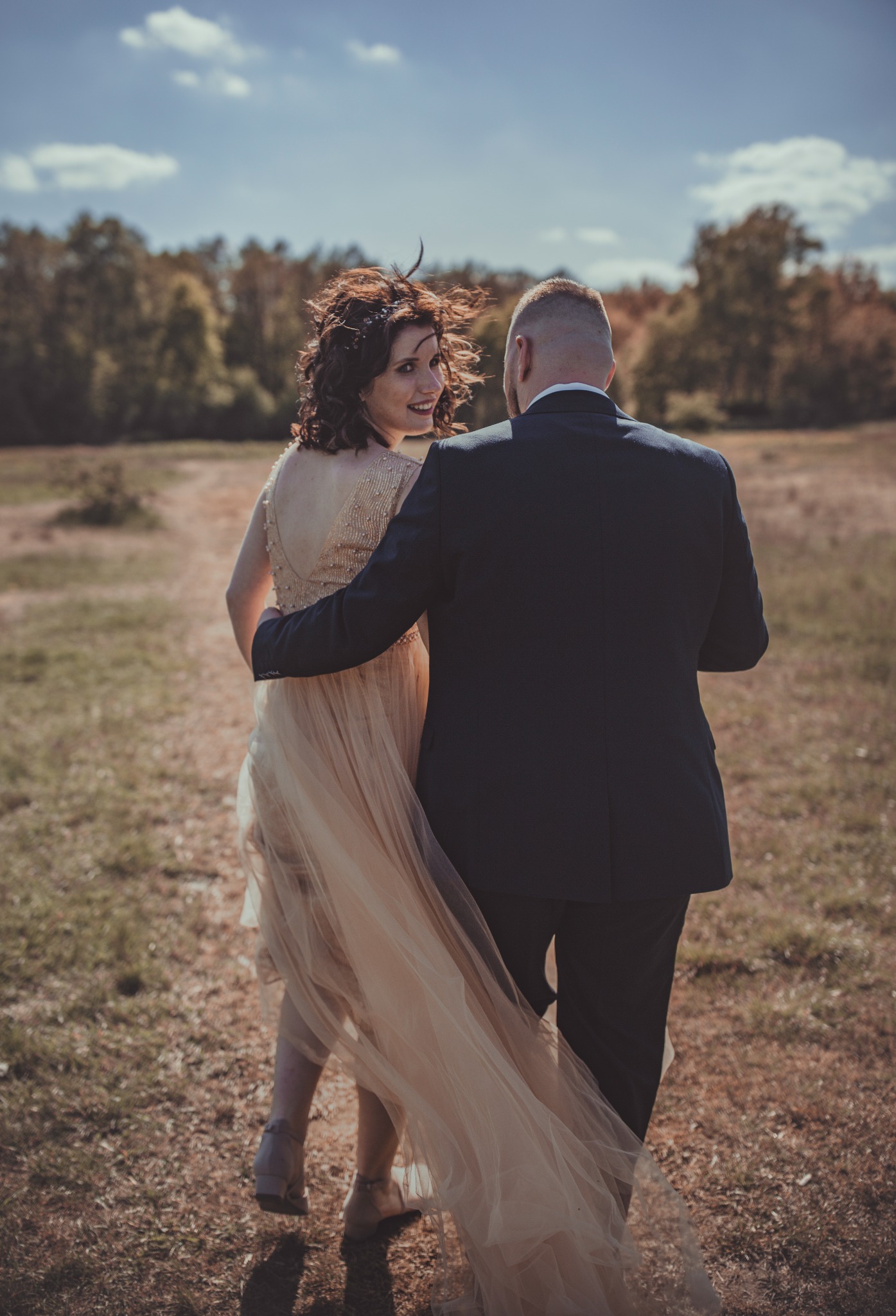 Para młoda idzie polną drogą, kobieta w sukni z odkrytymi plecami spogląda przez ramię, włosy rozwiane na wietrze, mężczyzna w ciemnym garniturze obejmuje ją ramieniem.