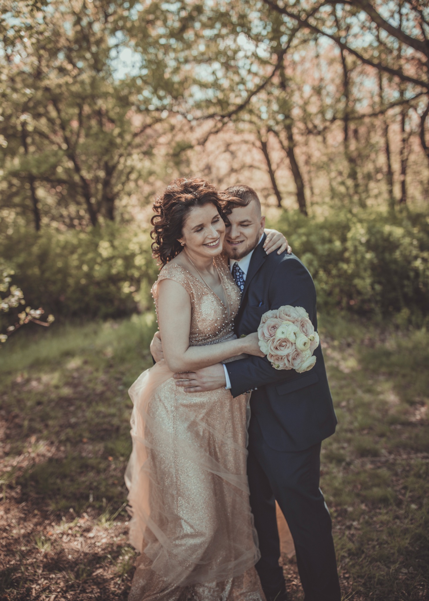 Uśmiechnięta para narzeczonych w objęciach na tle leśnej scenerii, panna młoda w złotej sukni, pan młody w granatowym garniturze z bukietem róż.
