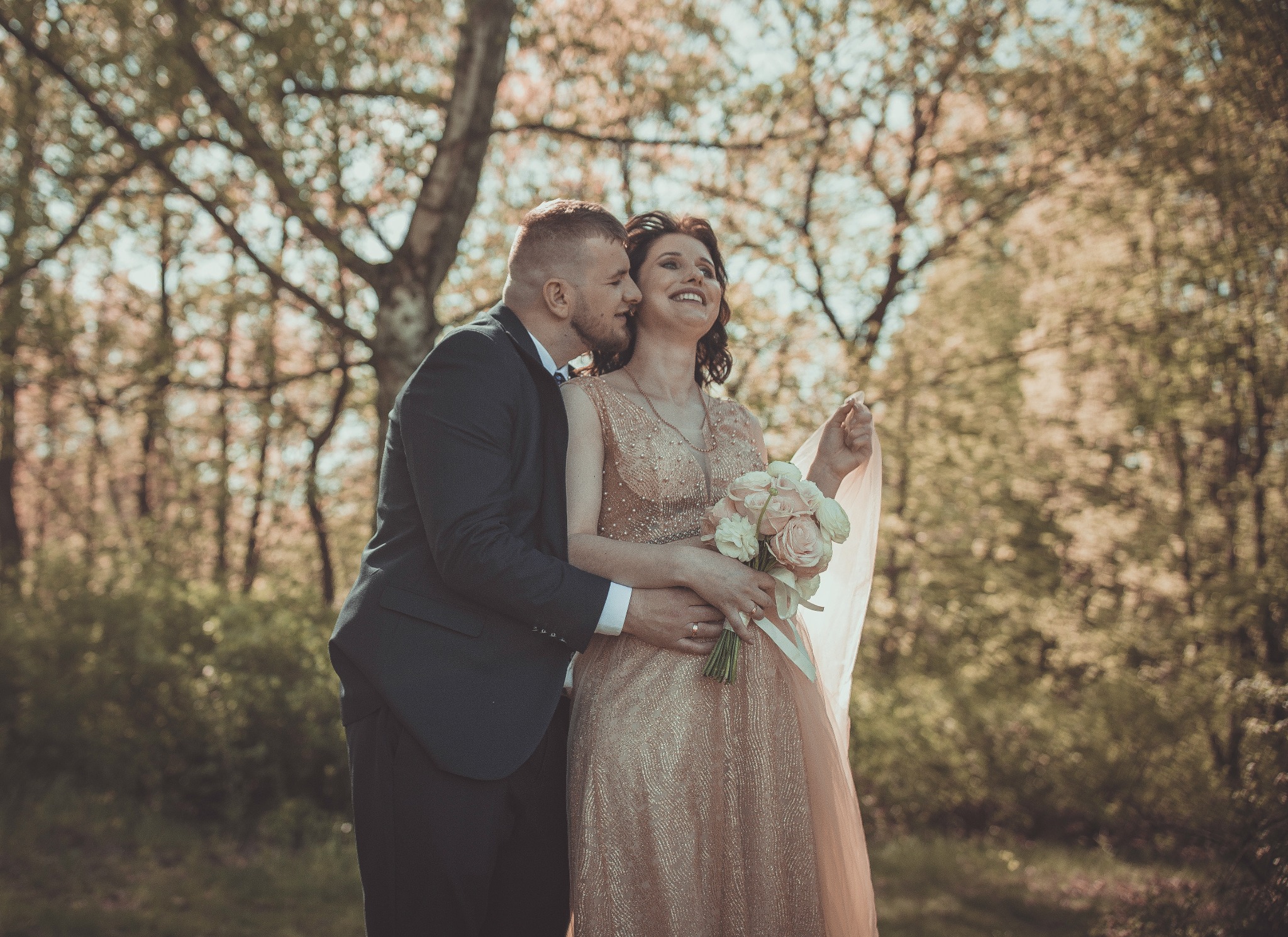 Uśmiechnięta para narzeczonych w plenerze: mężczyzna w granatowym garniturze obejmuje kobietę w złotej sukni, trzymającą bukiet różowych róż; w tle drzewa z zielonymi liśćmi.