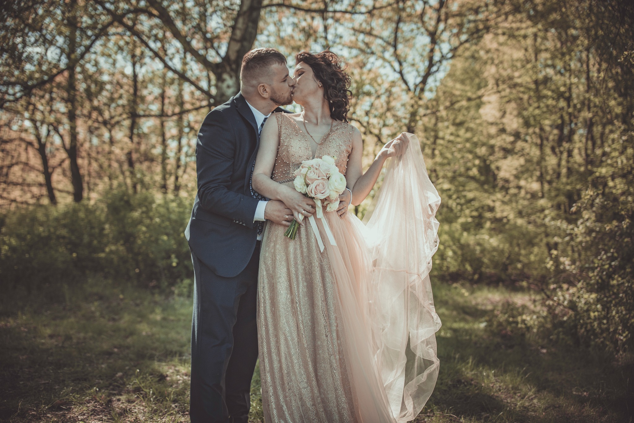 Para narzeczonych całuje się na tle leśnej scenerii, panna młoda trzyma bukiet różowych róż i zwiewną suknię w kolorze pudrowego różu i złota.