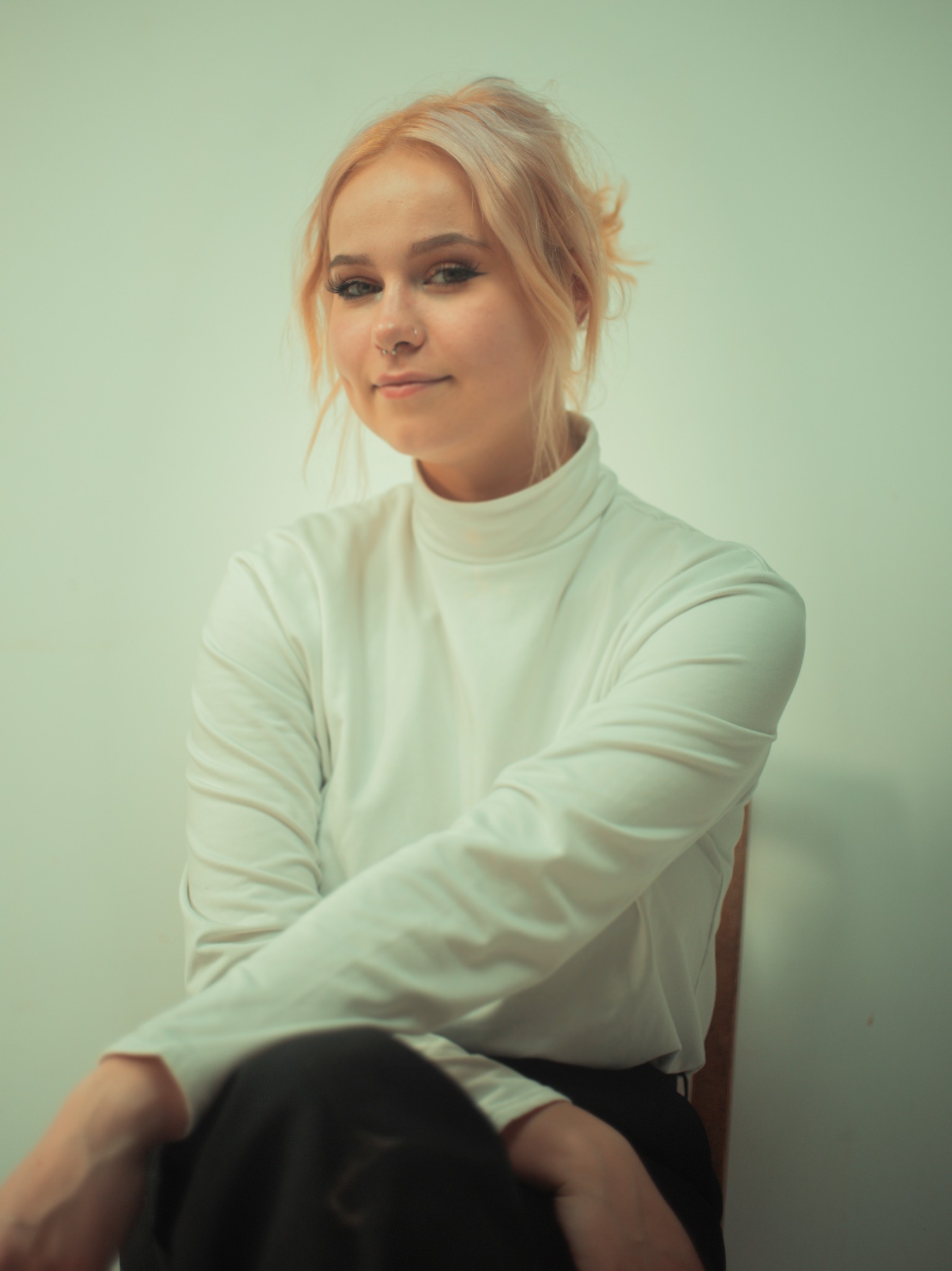 Portret młodej kobiety o jasnych włosach, ubrana w biały golf, siedzącej na krześle na tle jasnej ściany, z delikatnym uśmiechem i piercingiem w nosie.