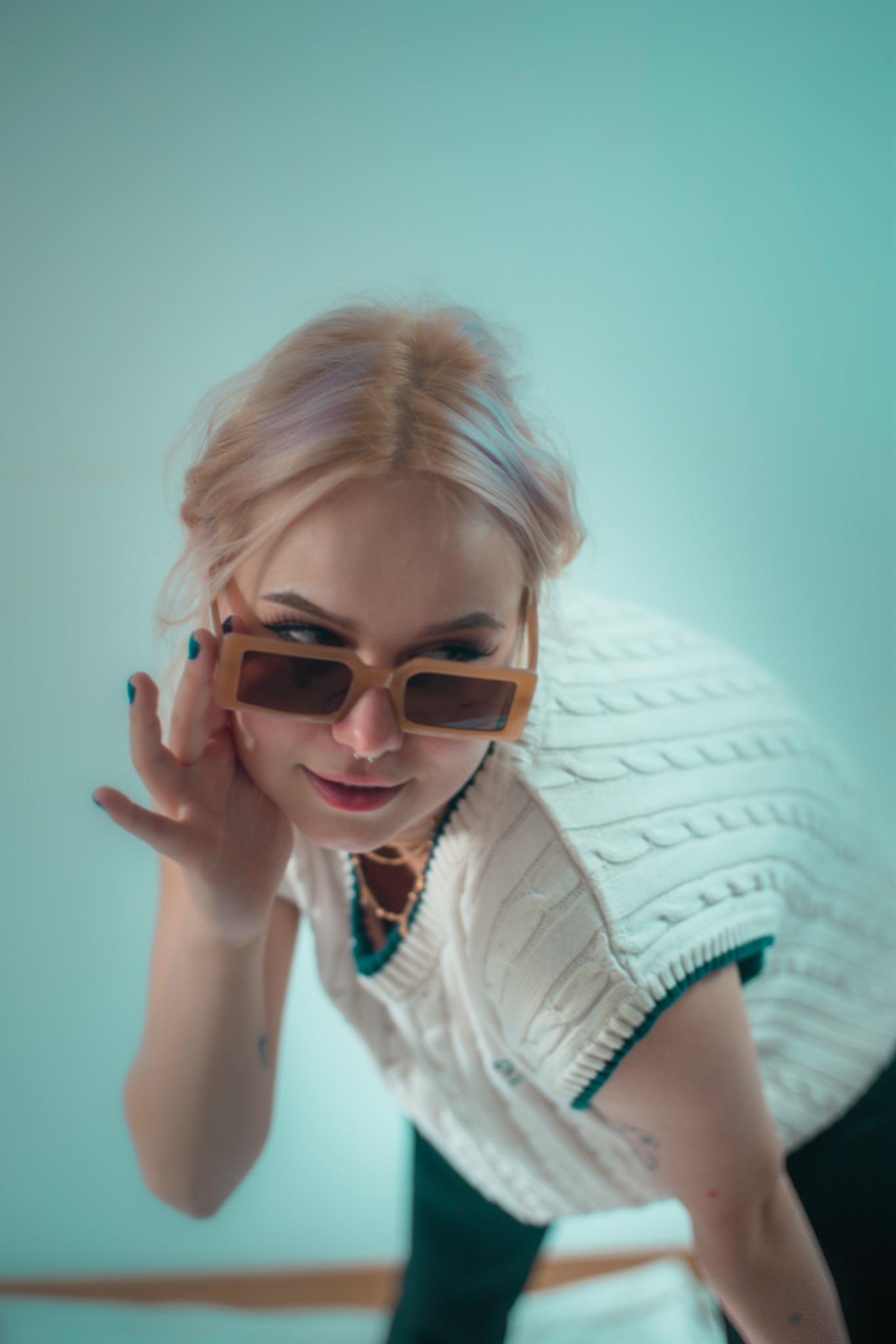 Młoda kobieta z blond włosami w okularach o grubych, beżowych oprawkach, ubrana w biały sweter z warkoczowym splotem, pozuje na rozmytym, jasnoniebieskim tle.
