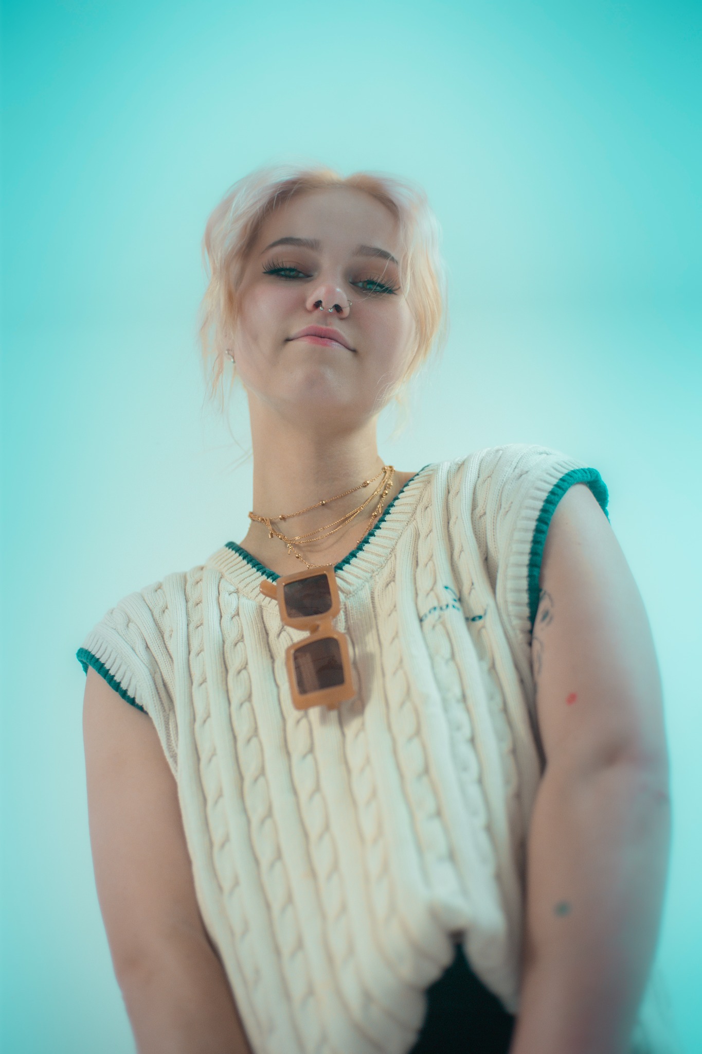 Młoda kobieta z blond włosami patrzy z góry, nosi sweter z dekoltem w serek, okulary przeciwsłoneczne na szyi i złote naszyjniki na tle turkusowego tła.