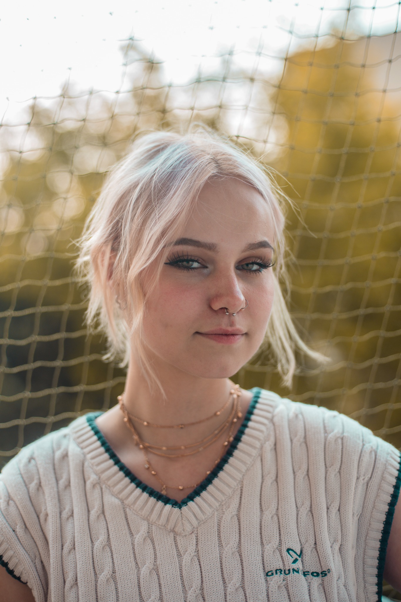Portret młodej kobiety z platynowymi włosami, piercingiem w nosie i delikatnym makijażem, ubrana w sweter w warkocze z logo firmy na tle rozmytej siatki.